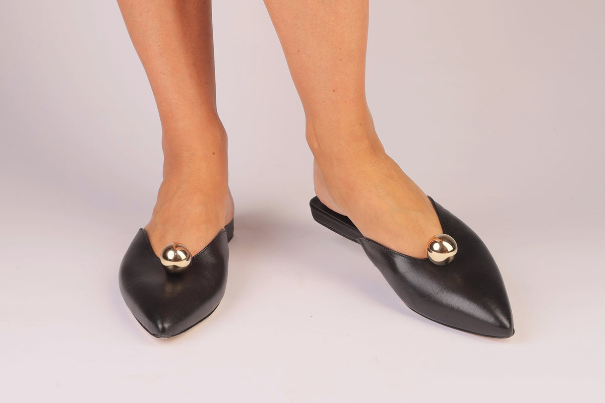 Black pointed-toe shoes with gold accents worn by a person on a light gray background