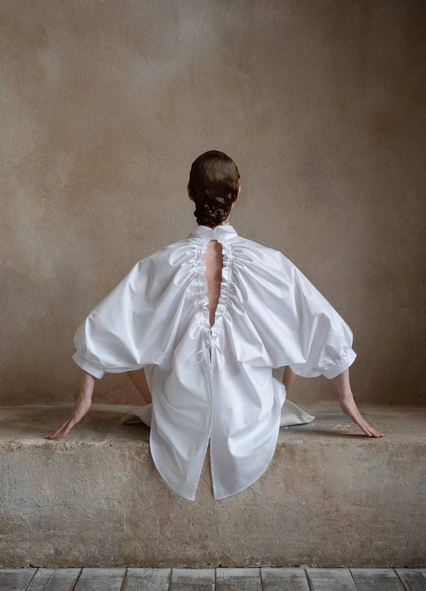 Person wearing a white blouse with open back sitting on a ledge against a plain wall.