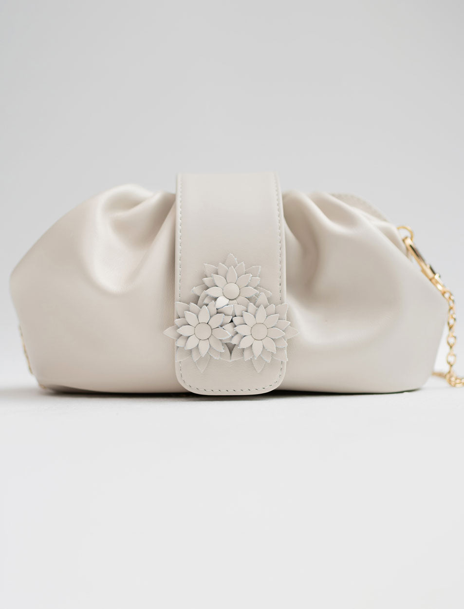 A cream-colored faux leather clutch bag with a gathered, slouchy silhouette, featuring a central strap adorned with a 3D floral embellishment and a gold chain strap visible on the right side, set against a light gray studio background.