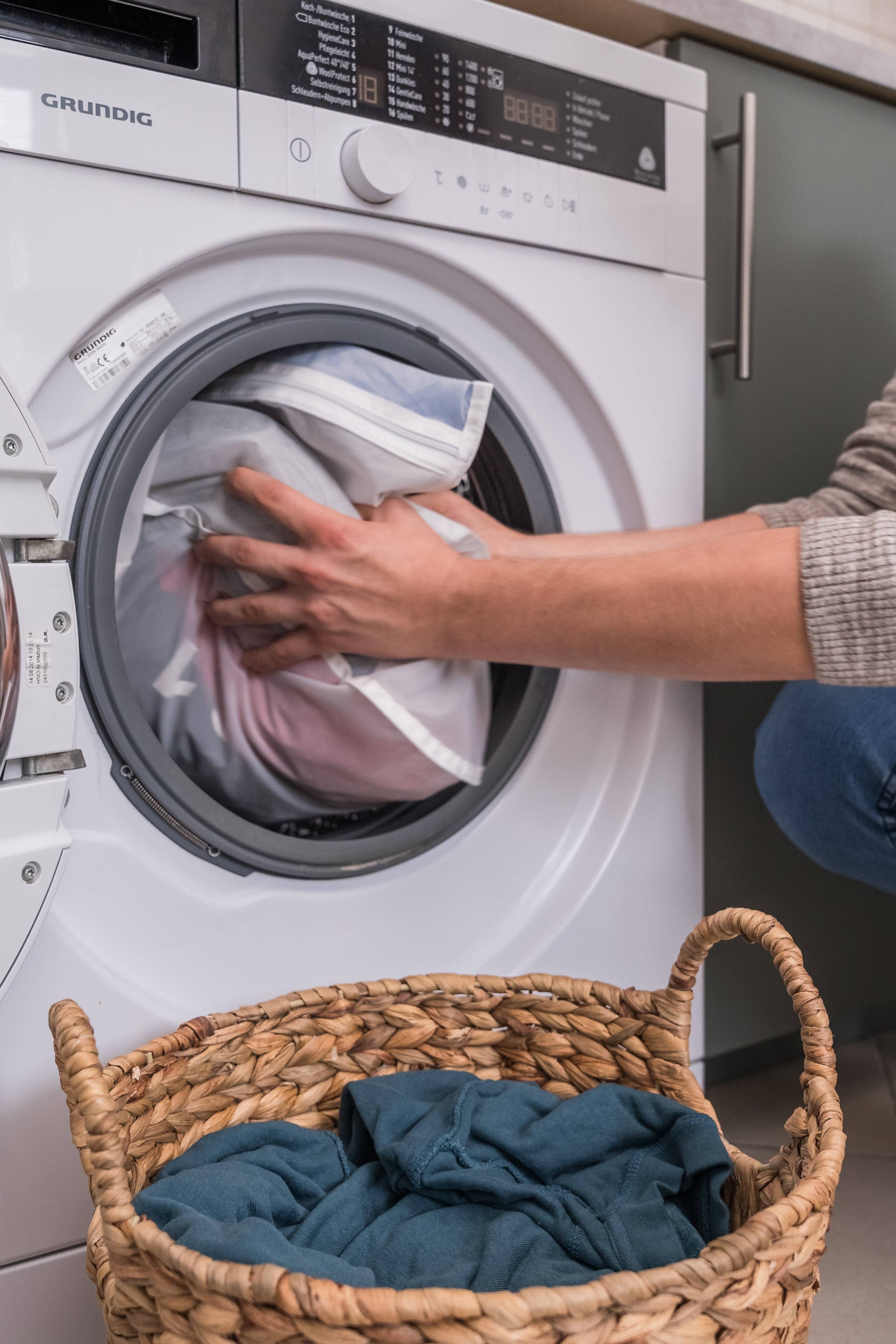 Person loading laundry into a washing machine with a basket of clothes nearby.