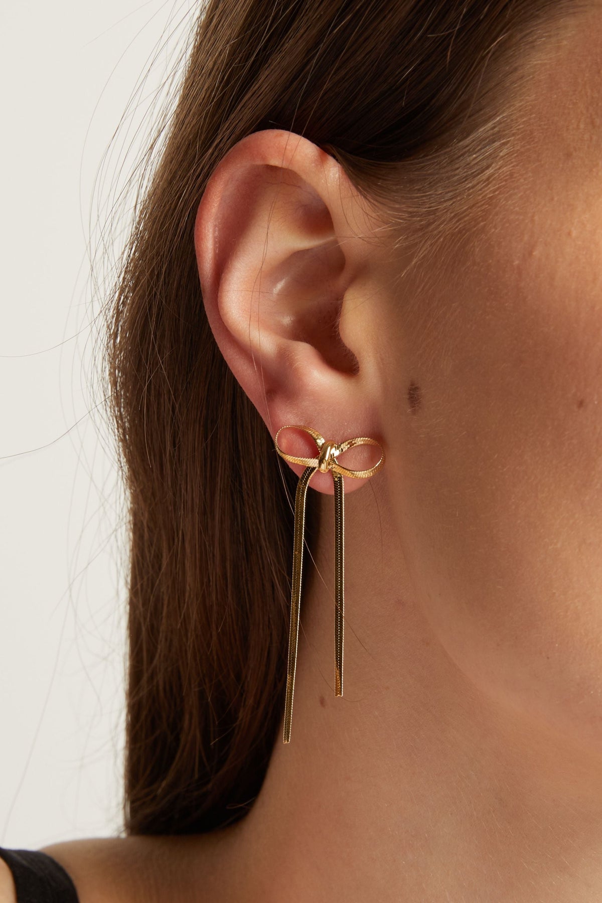 A close-up of a model's ear wearing a delicate gold dangle earring with a small bow detail and two long hanging bars.