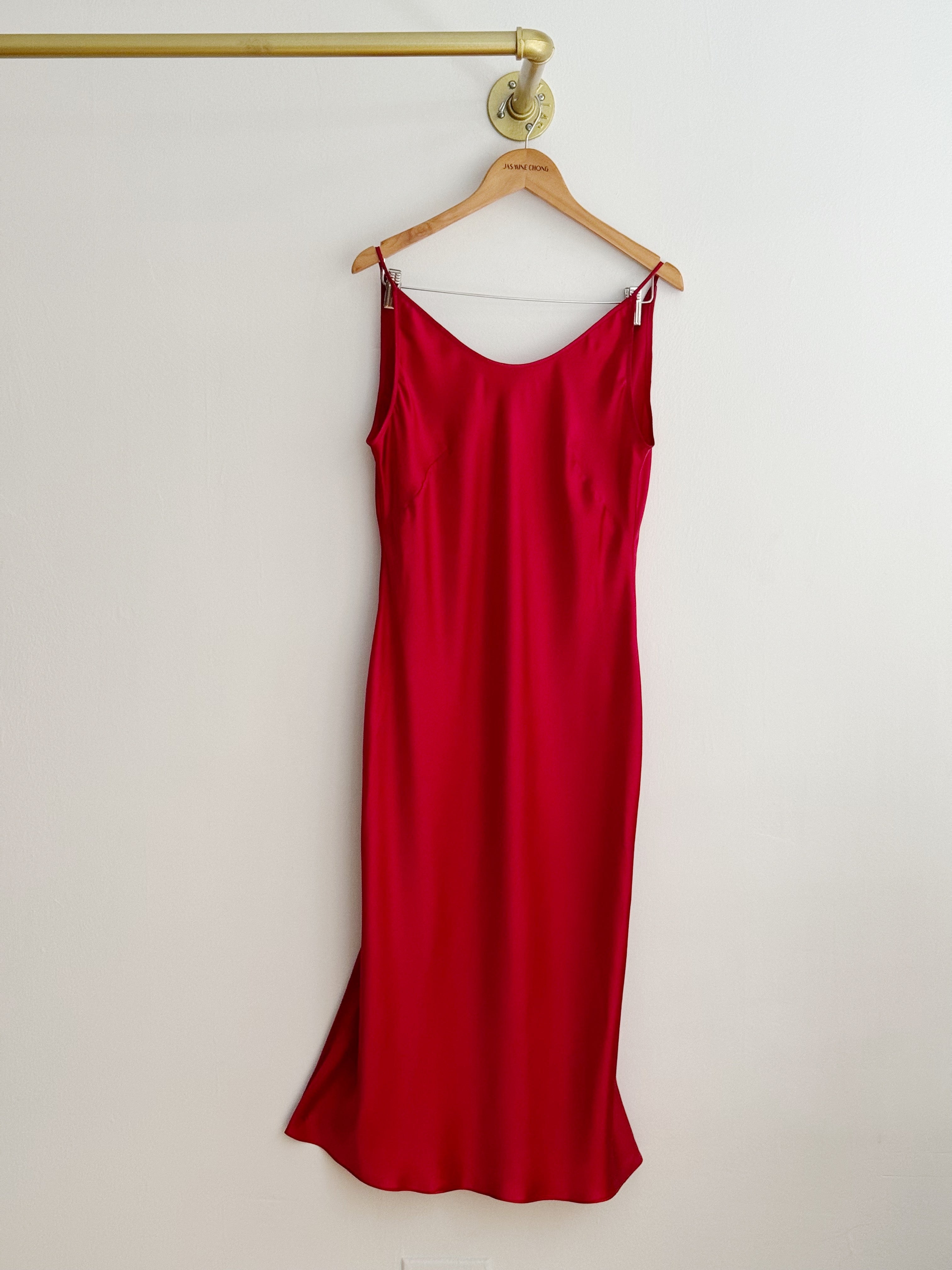 A red satin silk slip dress hanging on a wooden hanger from a gold wall rod against a plain off-white wall.