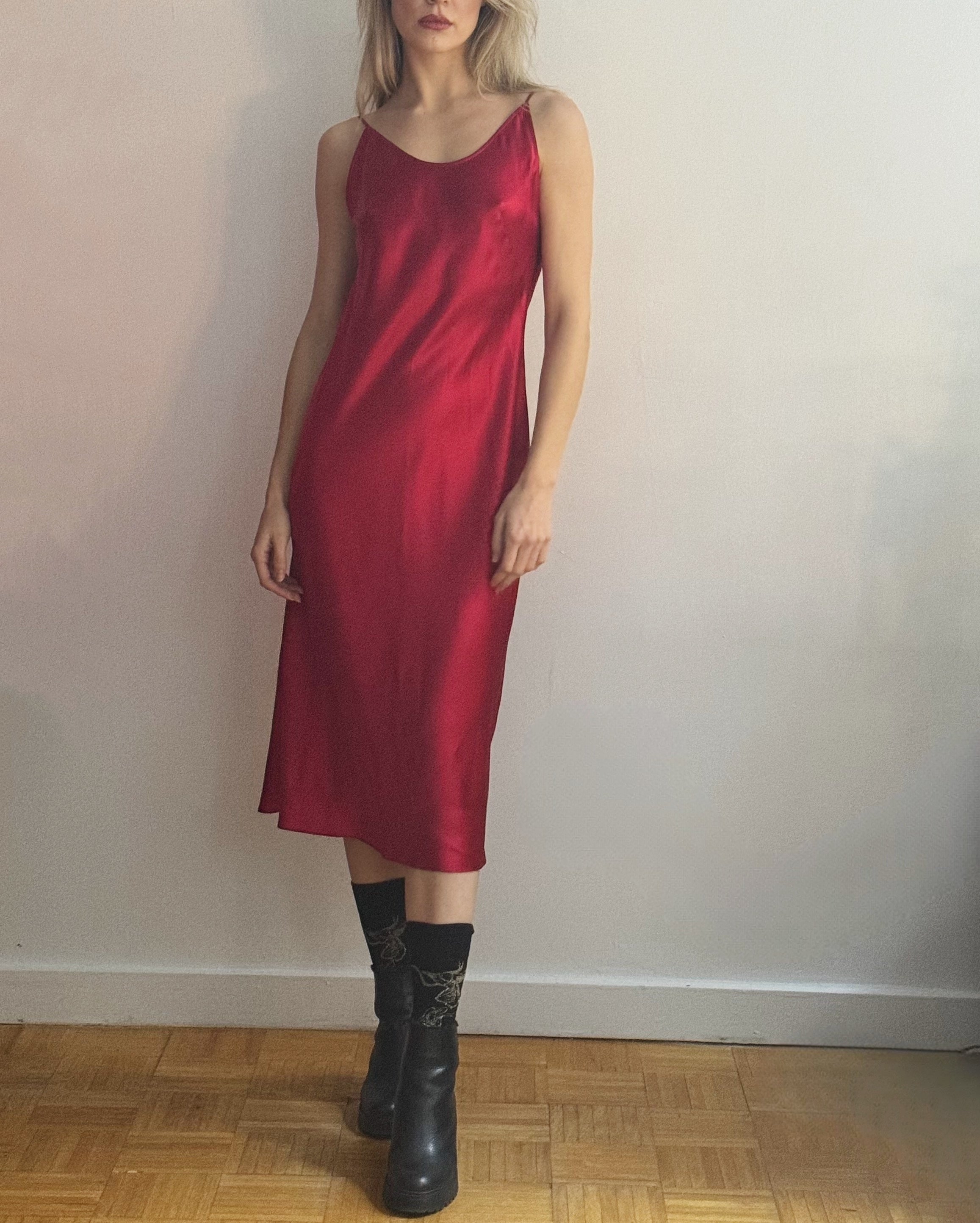 A woman wearing a shiny red satin slip dress with black ankle boots, standing against a plain off-white wall on a wooden parquet floor.