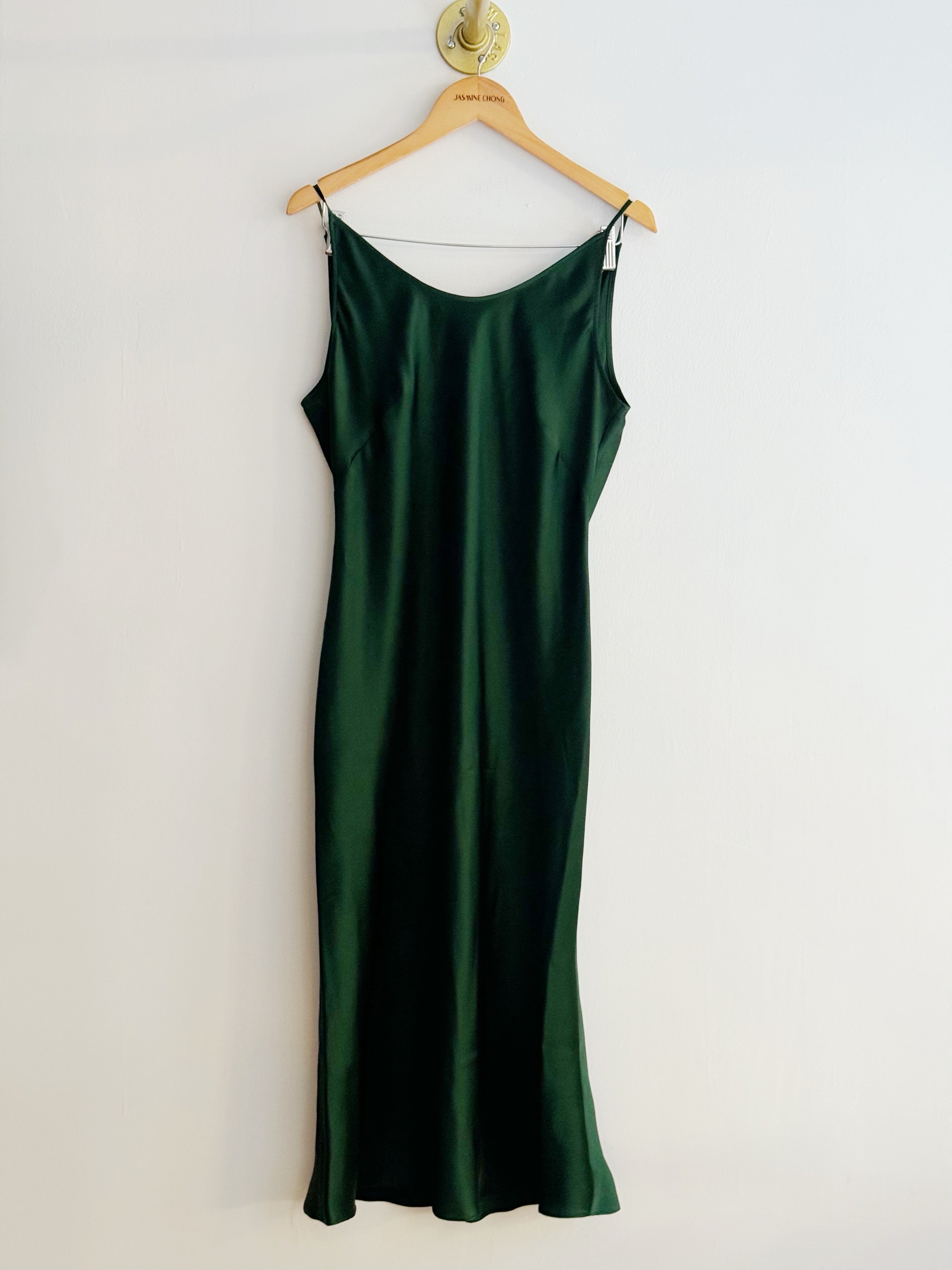 A dark green satin silk slip dress hanging on a wooden hanger against a plain white wall.