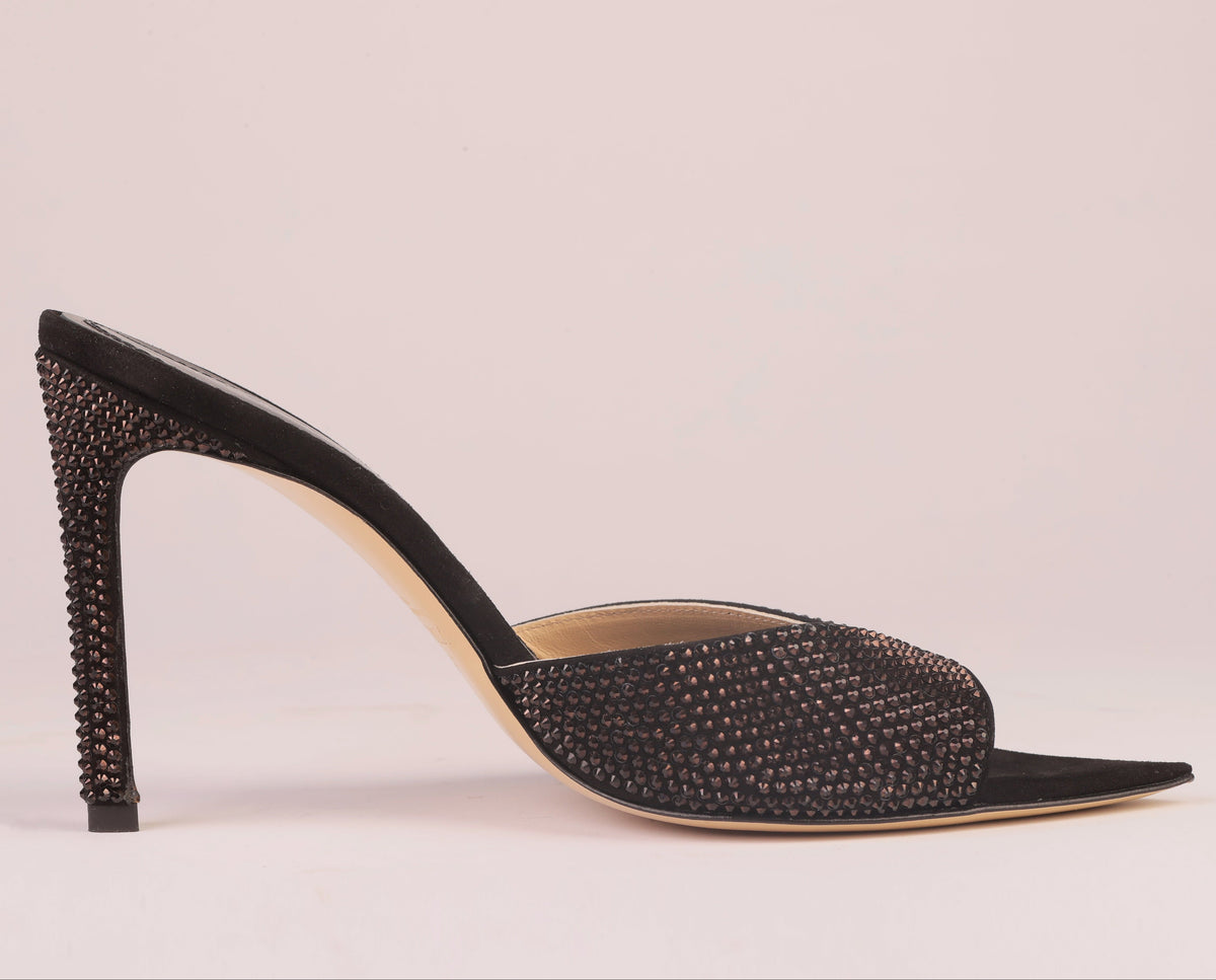 A single black pointed-toe mule high heel with a rhinestone-embellished upper and a studded column heel, shown in profile against a solid pale pink background.