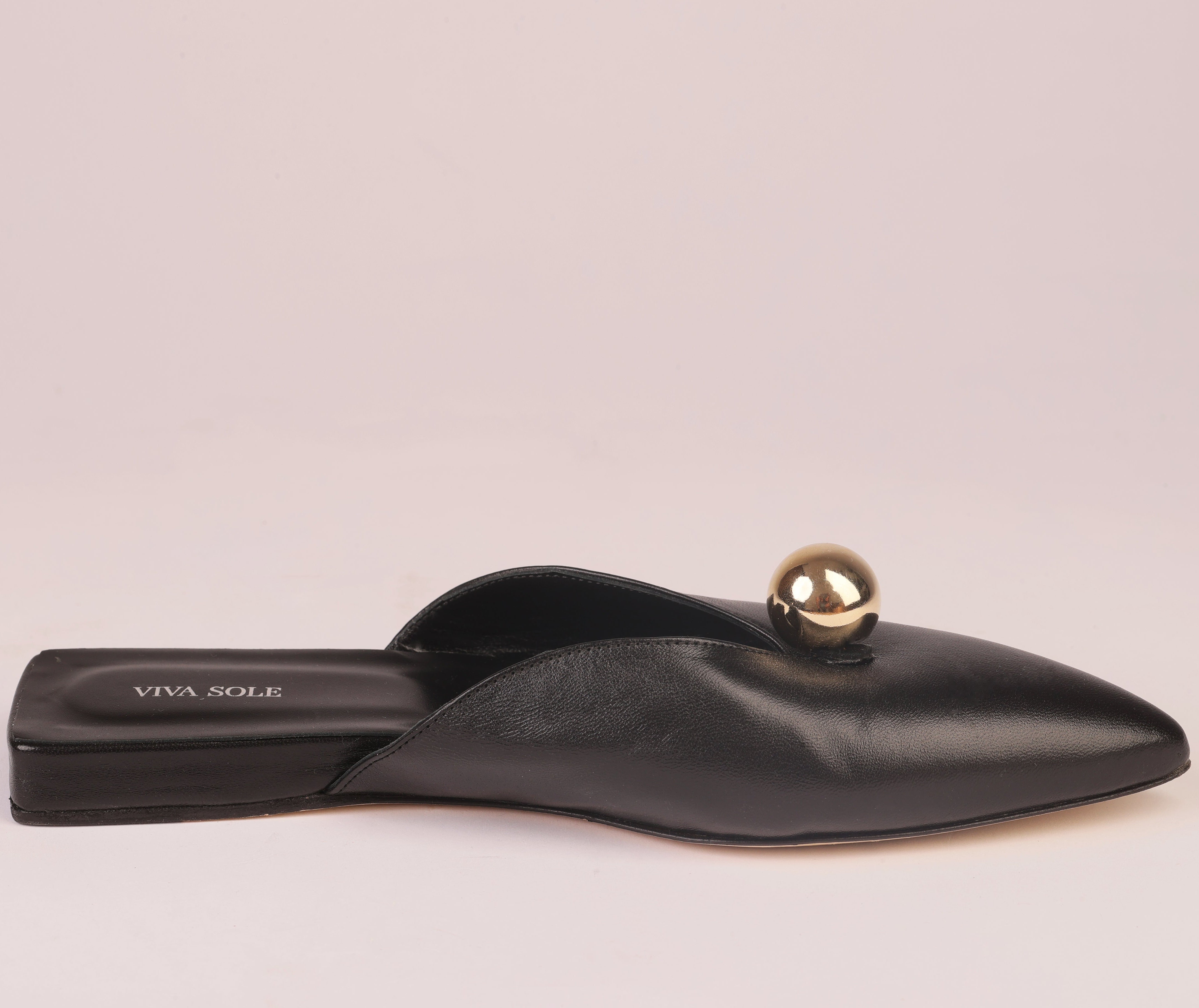 A black leather pointed-toe flat mule with a small metallic gold sphere detail on the strap, shown against a solid pale pink background. The brand name 'VIVA X OLE' is printed on the insole.
