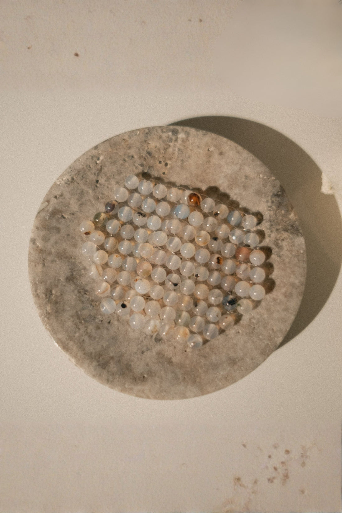 A round stone dish with a textured beige and gray surface, holding an agate stone eye stone eye compress made of small, round, milky white agate beads with subtle gray and brown speckles, photographed on a plain light beige background.