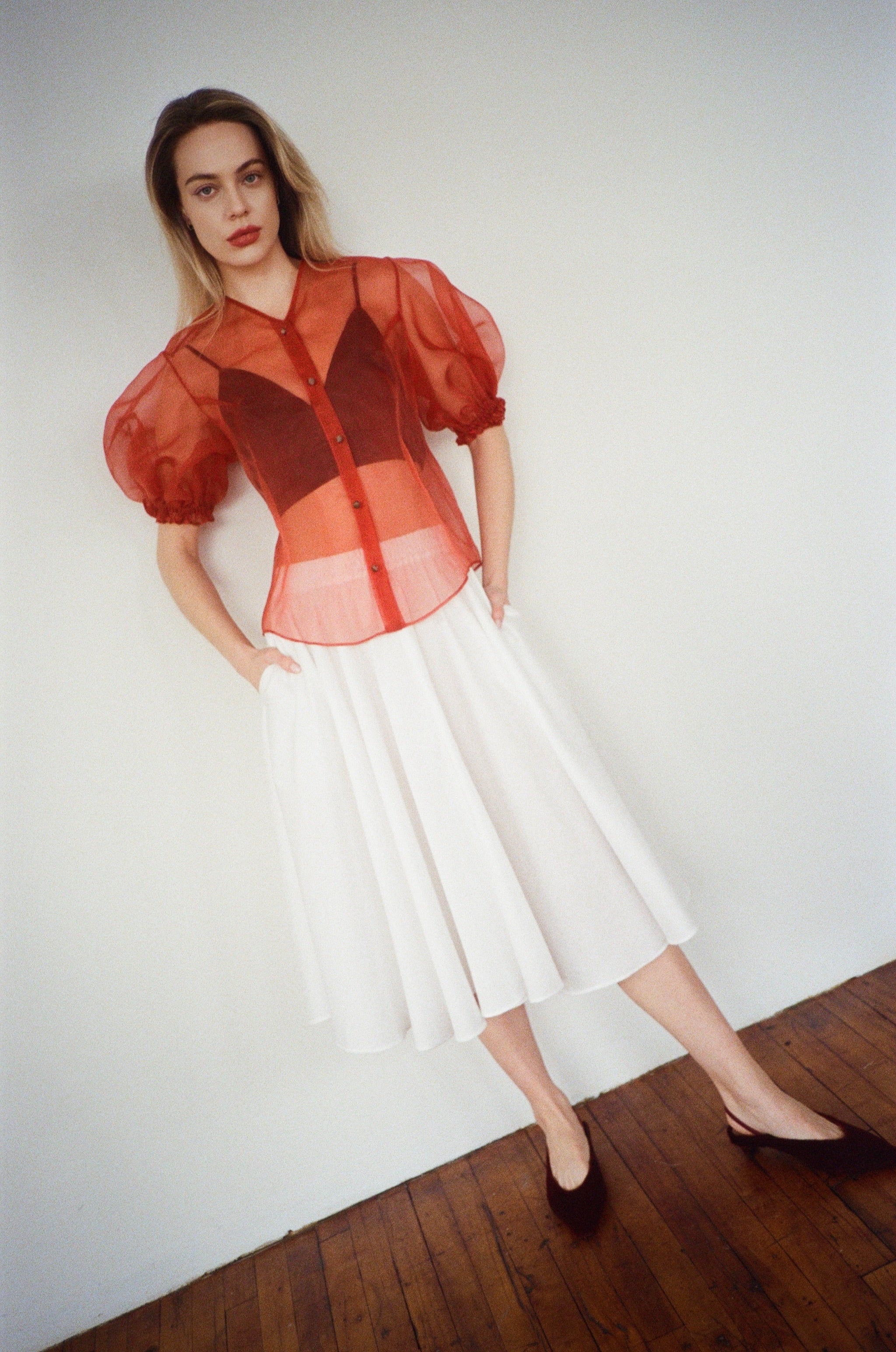 Woman wearing a red sheer silk blouse with puffed sleeves and a white cotton midi skirt against a plain background