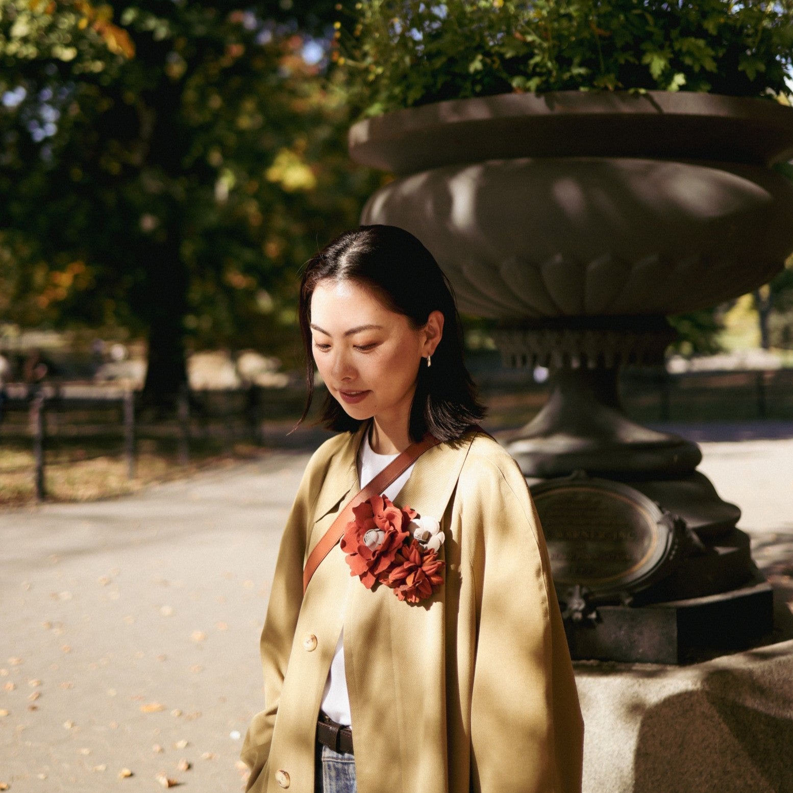 Woman in a beige coat with flower brooches standing in a park with a large stone vase in the background.