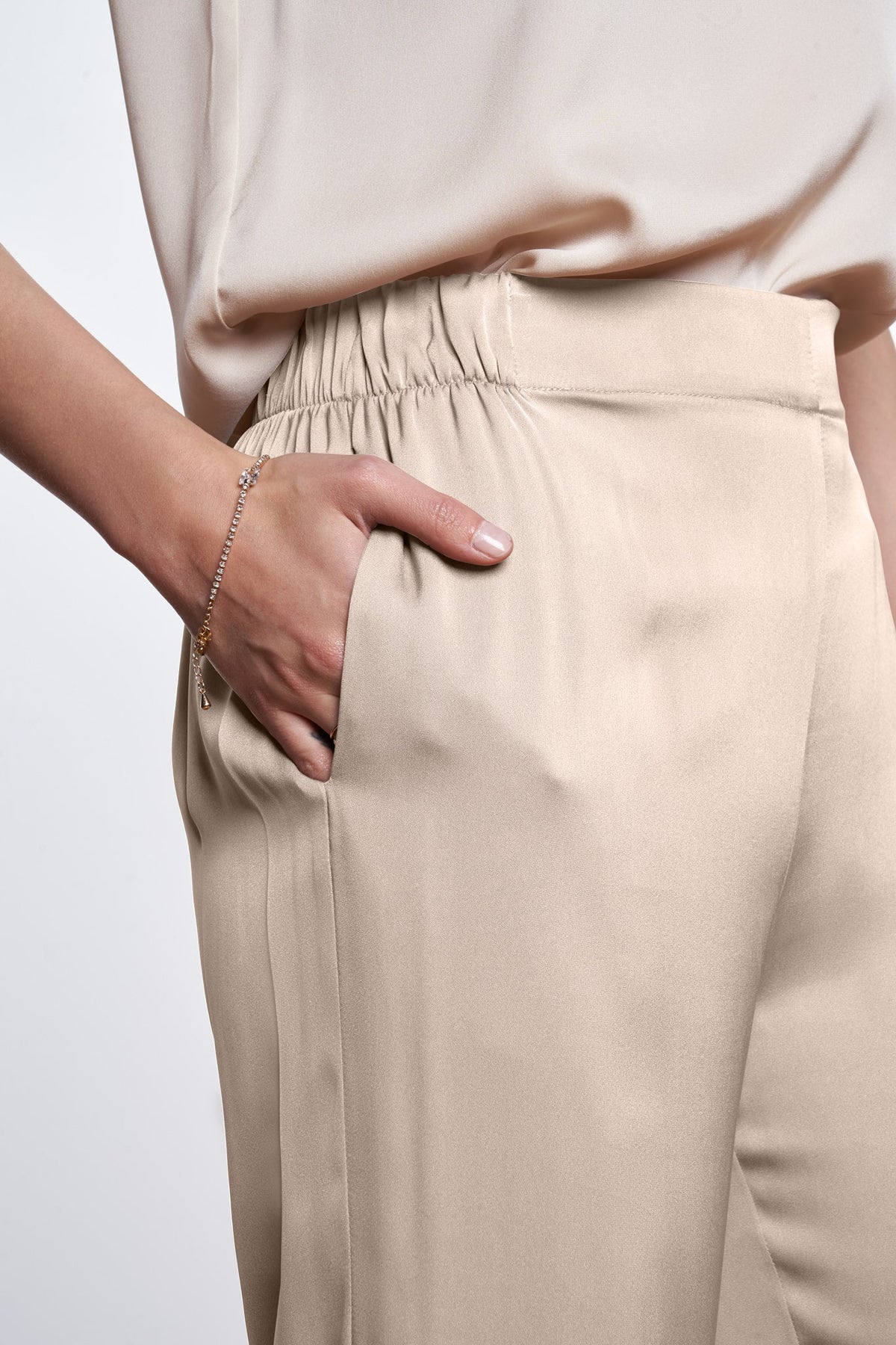 Close-up of a person wearing beige wide-leg silk trousers with an elastic waistband and side pockets, paired with a matching beige top against a light gray background.