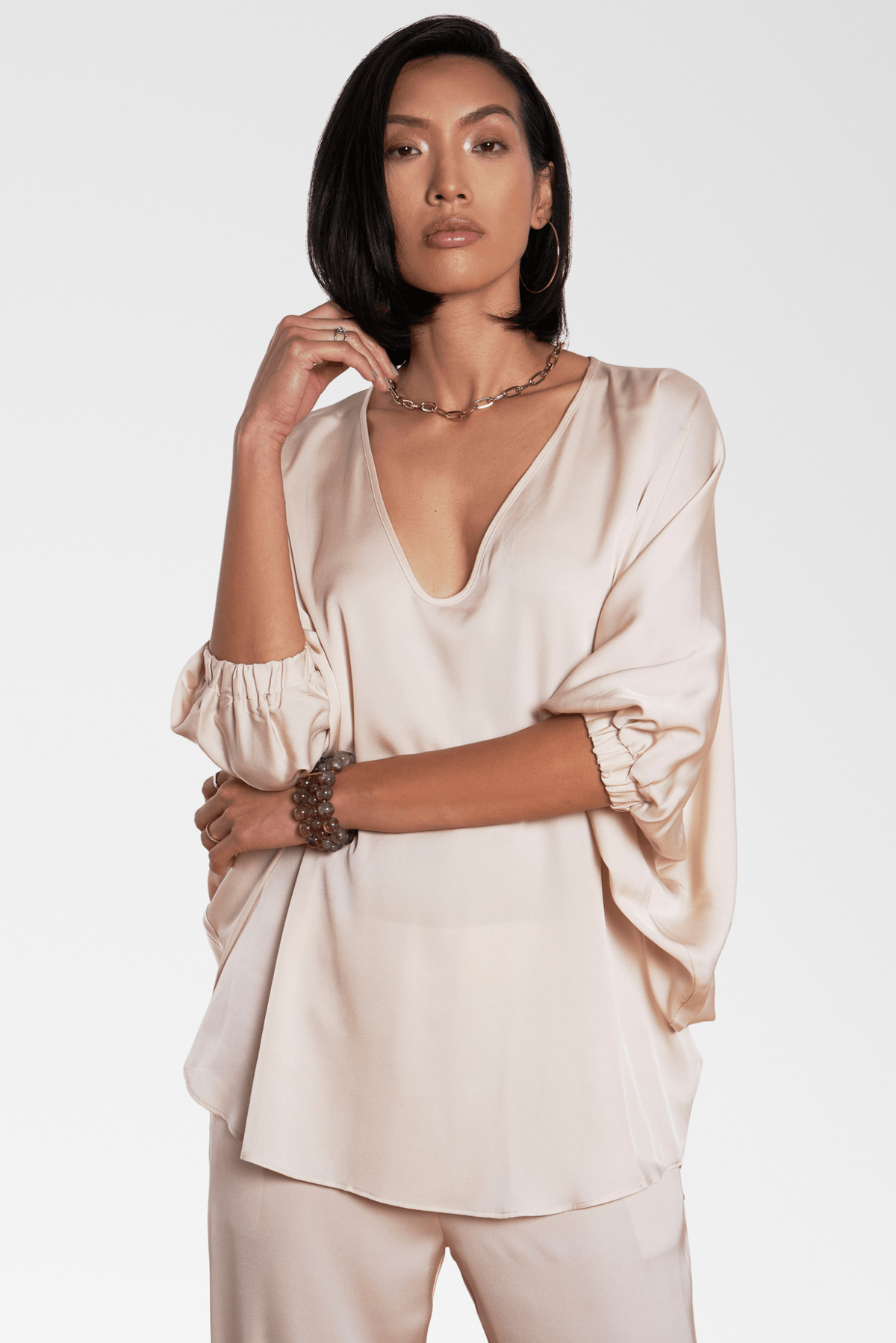 A model wearing a light beige satin silk blouse with a deep V-neck, gathered elbow-length sleeves, and matching beige trousers, set against a plain light gray studio background.