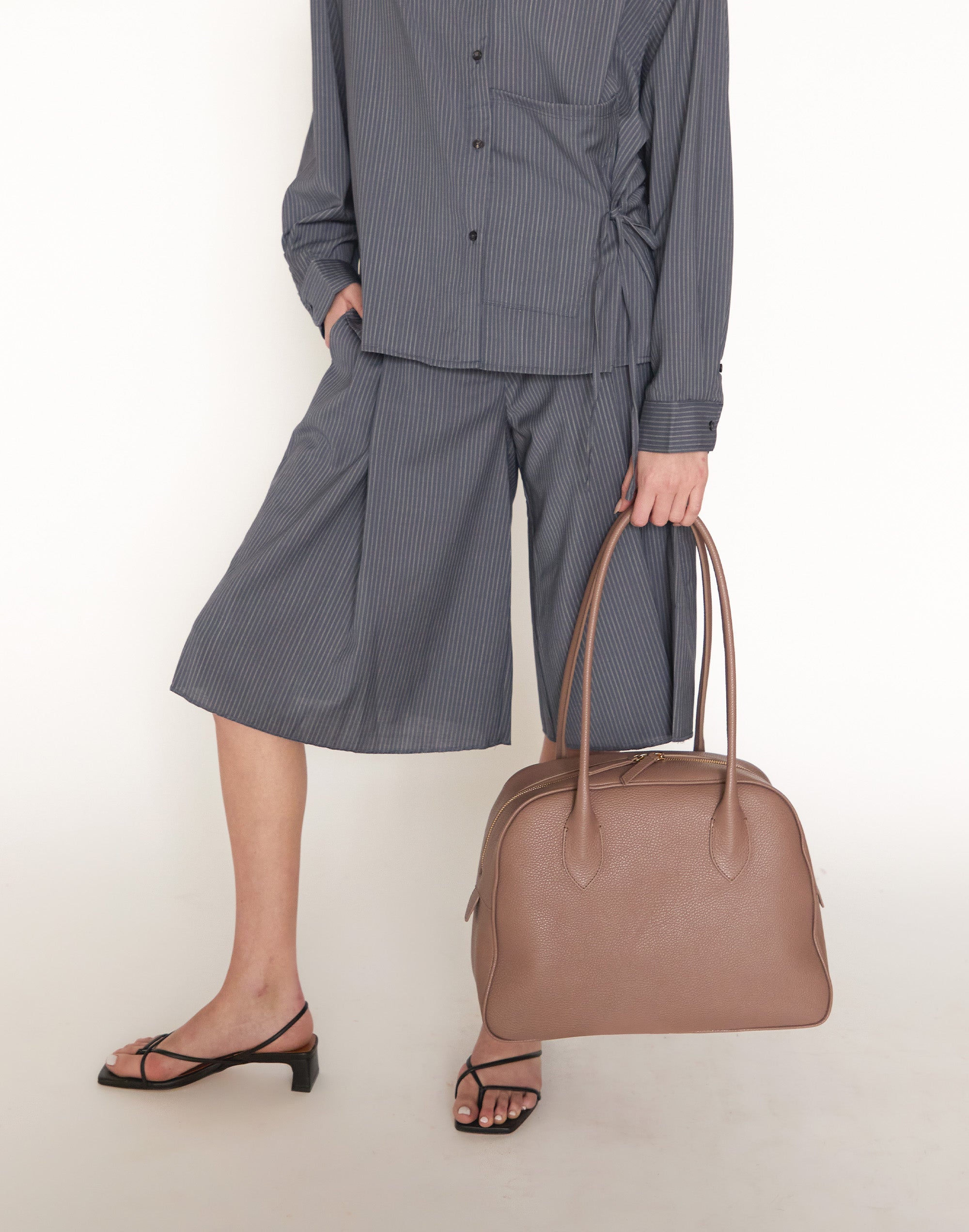 Person wearing a gray outfit with wide-leg pants holding a brown handbag on a white background