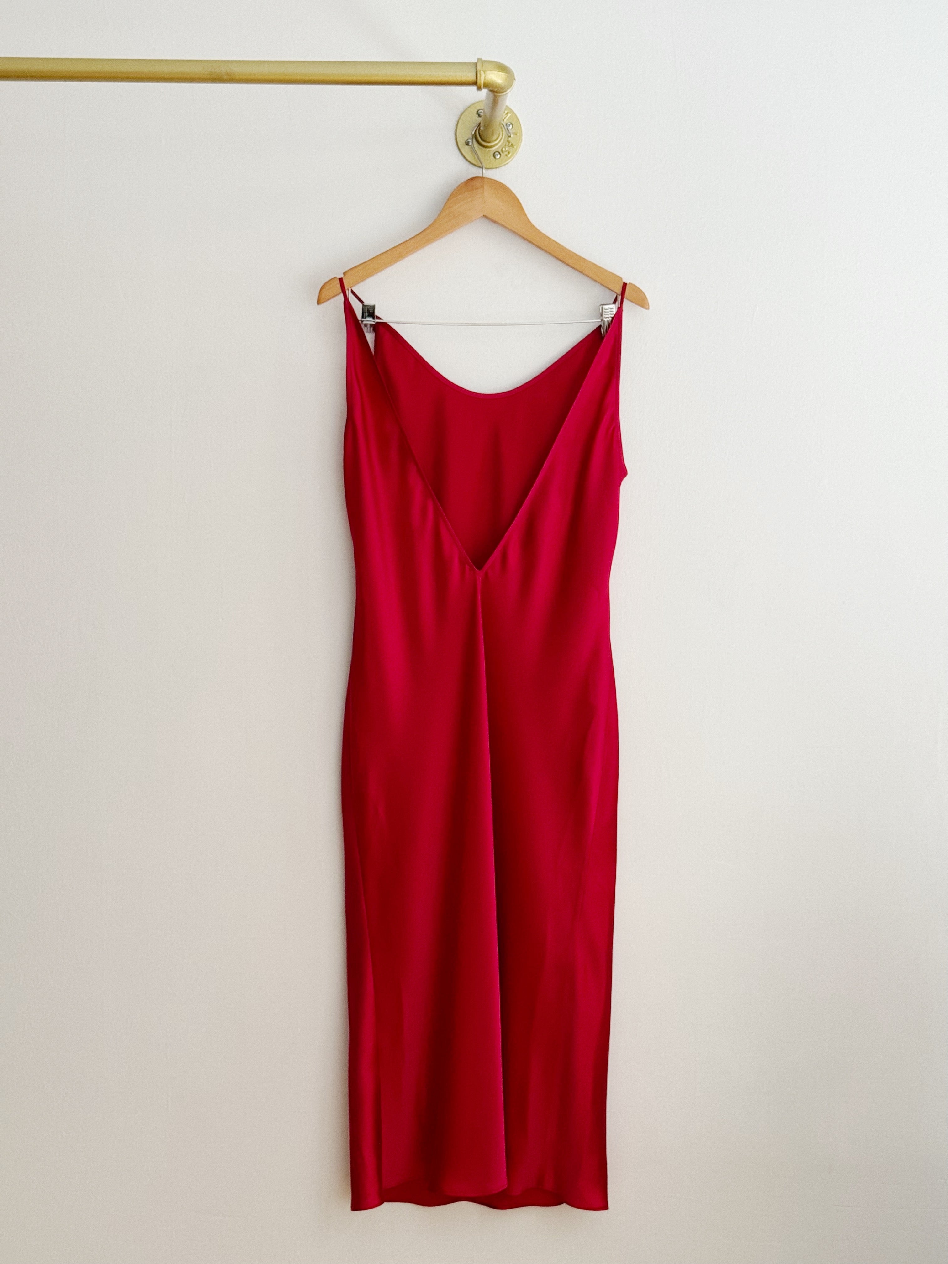 The back of a sleeveless red silk slip dress with a deep V-neckline hanging on a wooden hanger from a gold metal rod against a plain off-white wall.