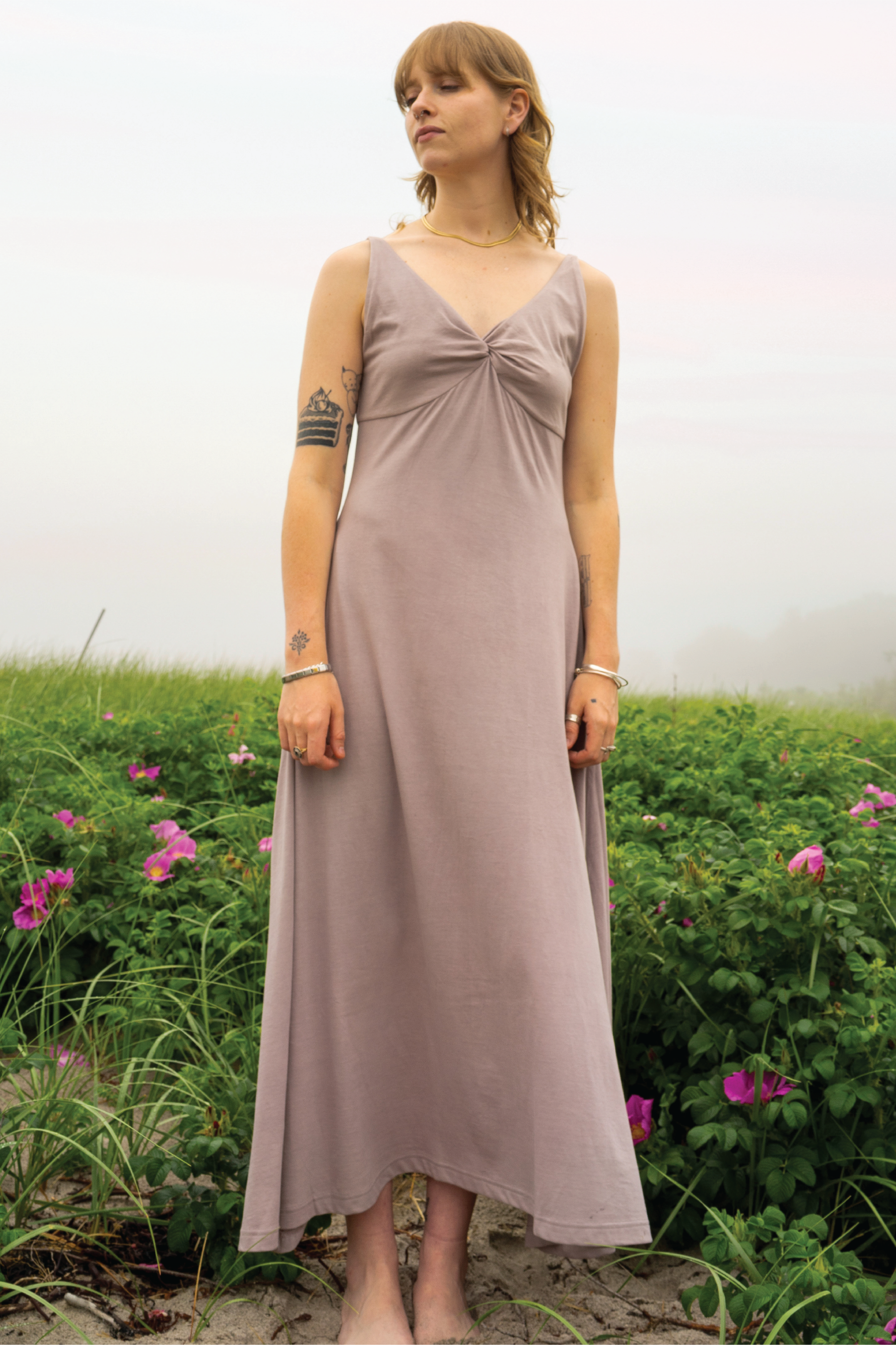 Woman in a long, light-colored dress standing in a field with greenery and flowers.