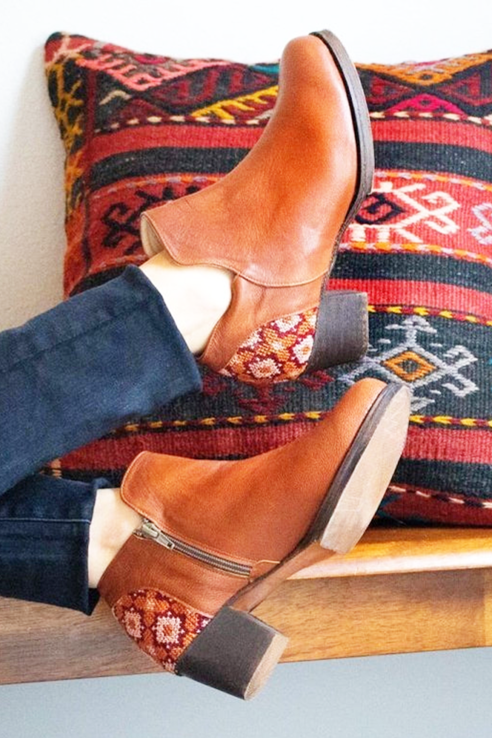 Brown leather boots on a person's feet with a colorful patterned pillow in the background