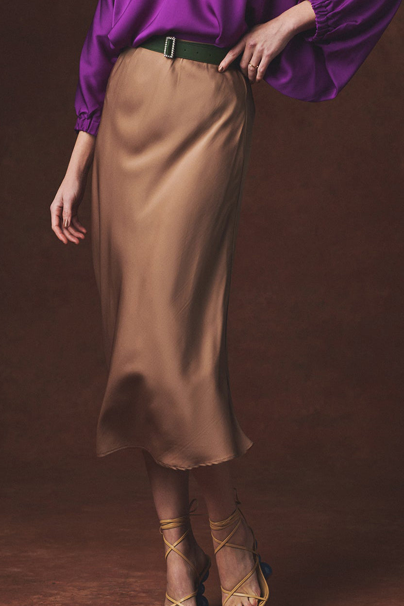 A model wearing a brown satin silk midi skirt with a dark green belt, paired with a purple blouse and strappy heels, set against a dark brown studio background.