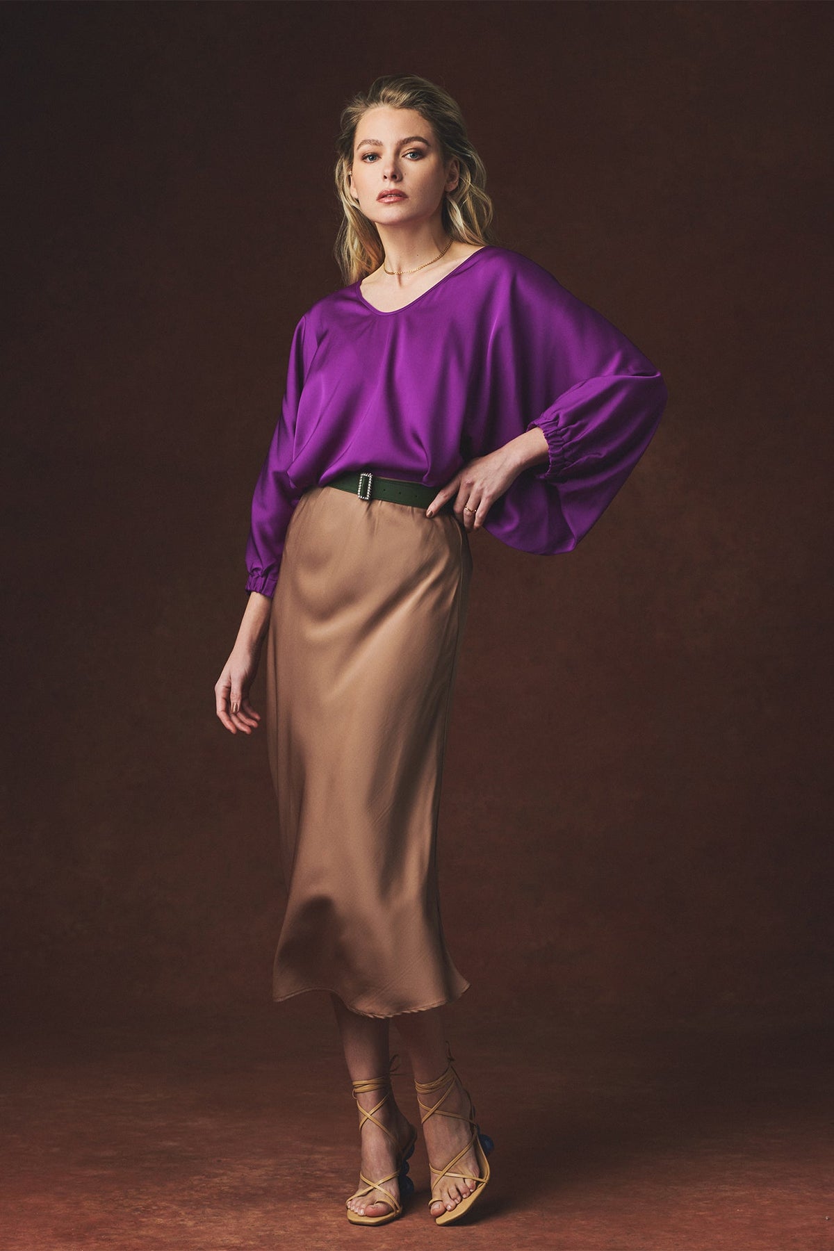 A woman models a purple long-sleeve blouse with voluminous balloon sleeves and a brown satin midi skirt, styled with a black belt and strappy heels against a dark brown studio backdrop.