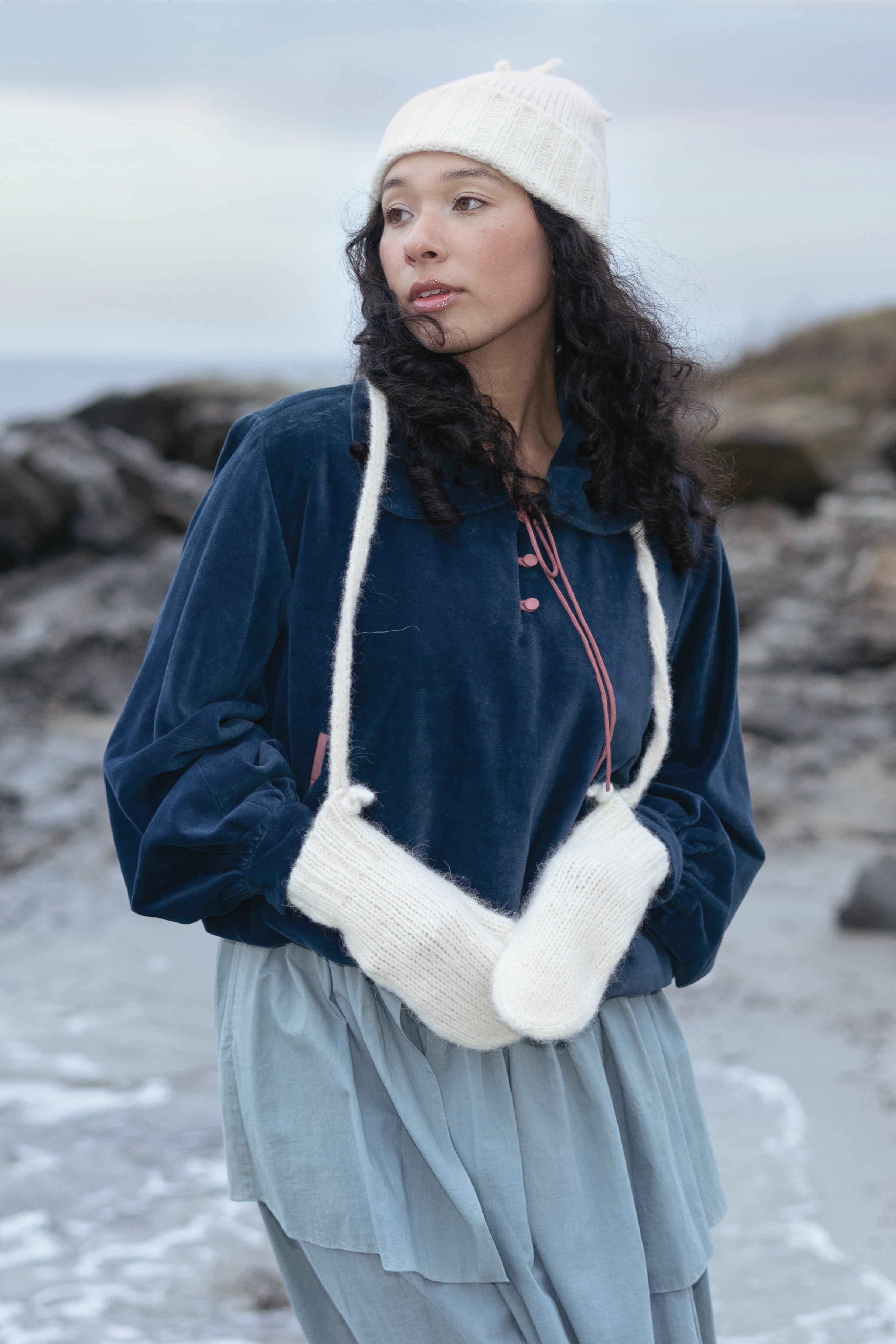 Woman wearing a blue jacket, white beanie, and gloves standing on a rocky beach.