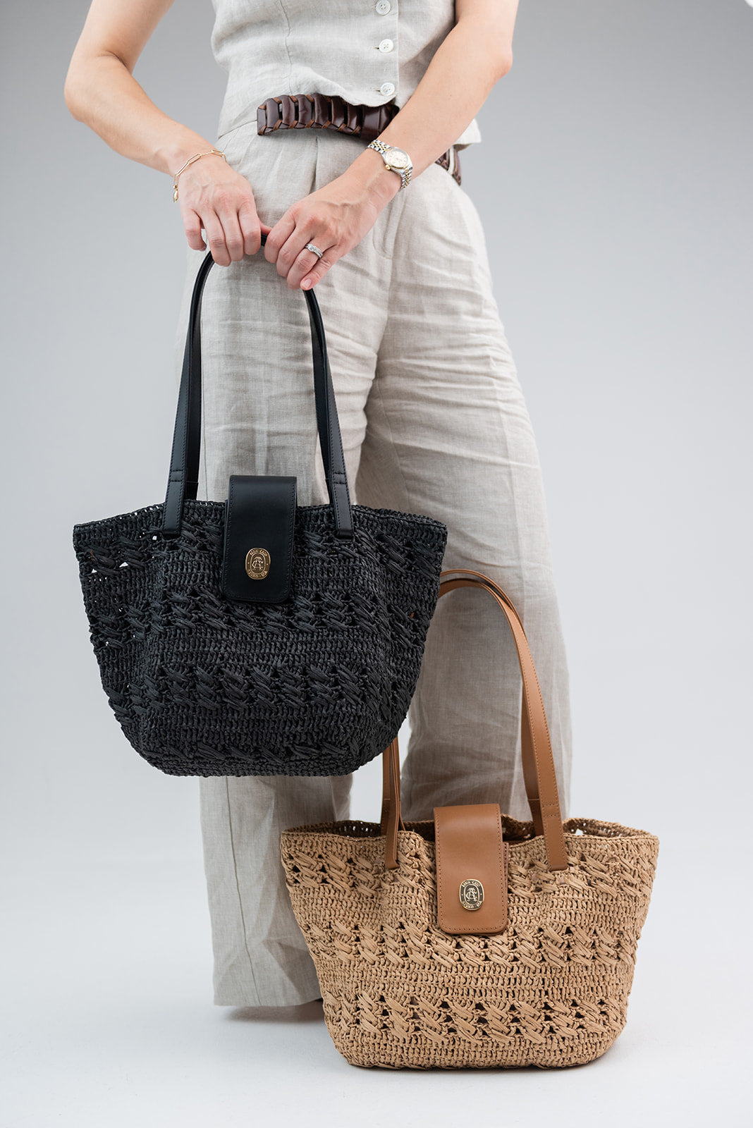 Two woven straw tote bags are shown against a light gray studio background. The top bag is black with black handles and a black front tab; the bottom bag is natural tan with tan handles and a tan front tab.