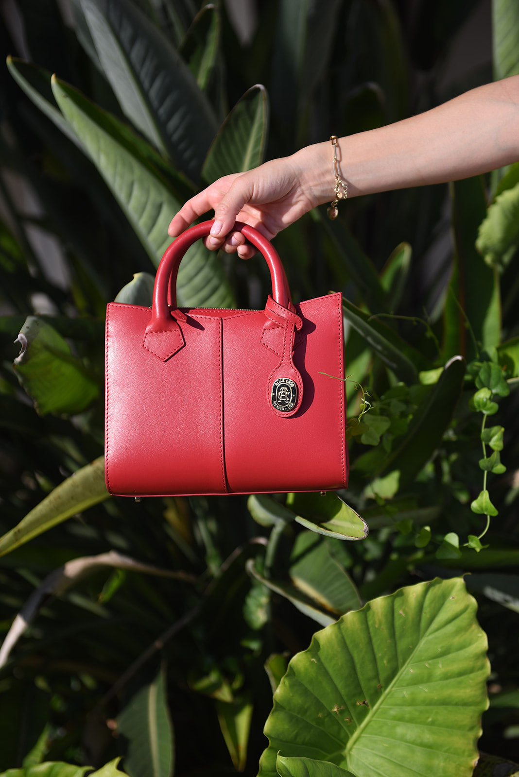 A red leather handbag with top handles and a gold-tone logo clasp, held by a person's hand against a background of lush green tropical leaves.