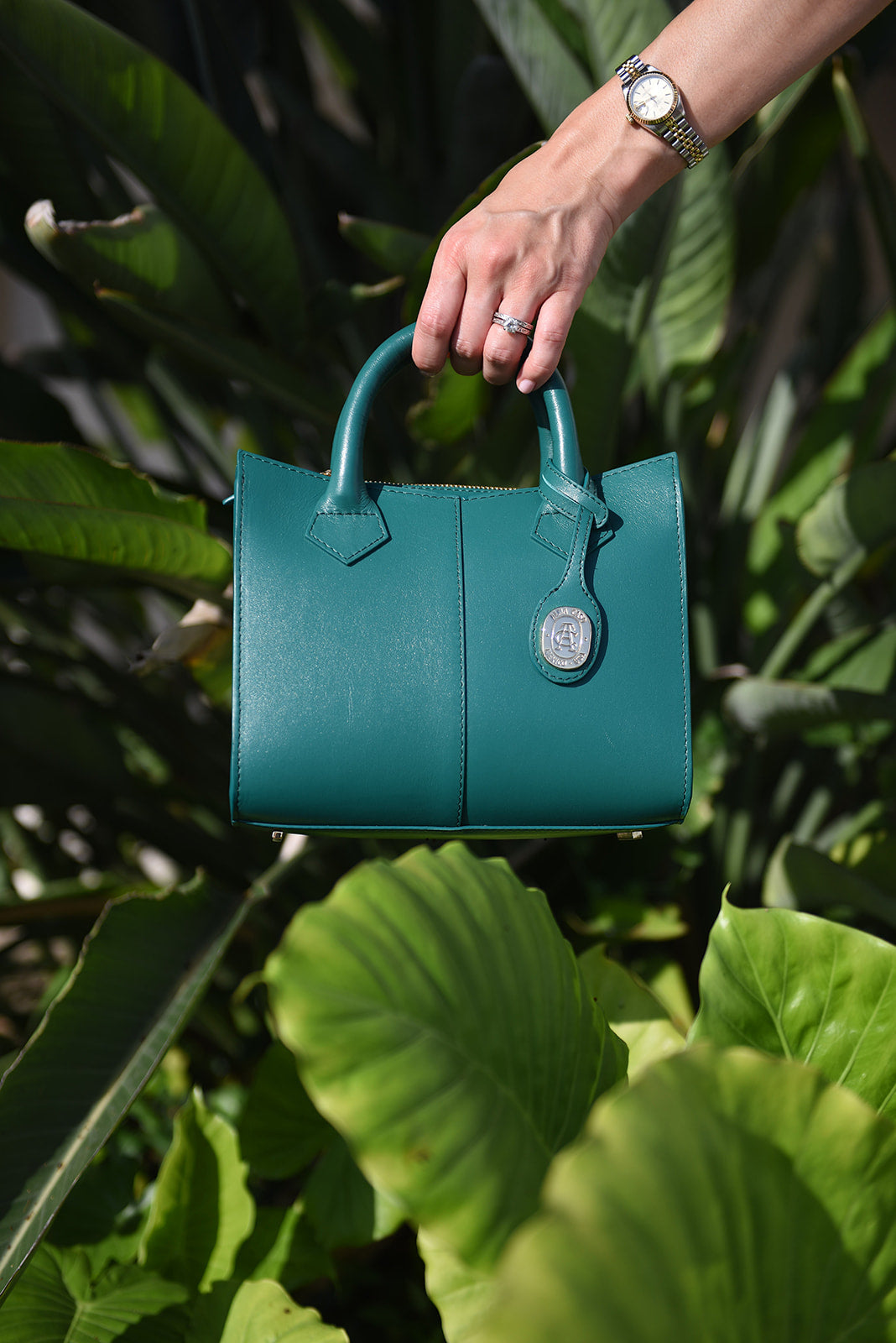 A teal leather handbag with top handles and a silver-tone circular charm, held by a person's hand against a background of lush green tropical leaves.