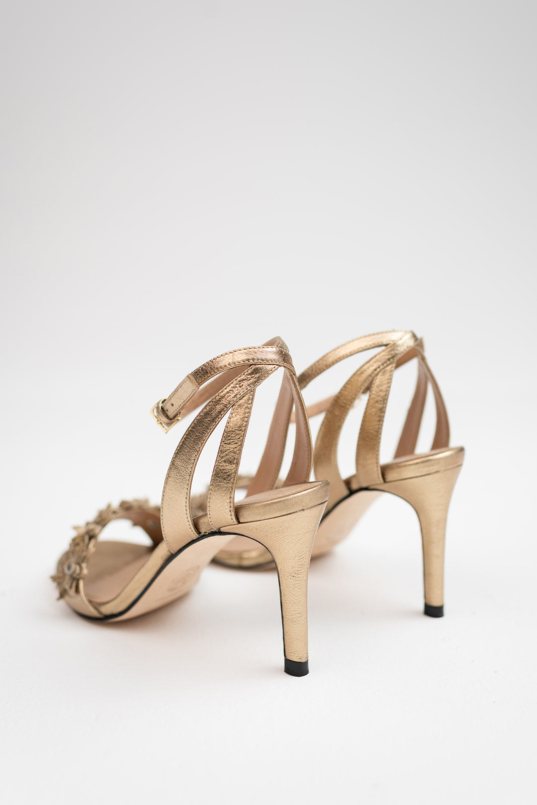 A pair of metallic gold strappy high-heeled sandals with a jeweled toe detail, photographed from a side-rear angle on a light gray studio background.
