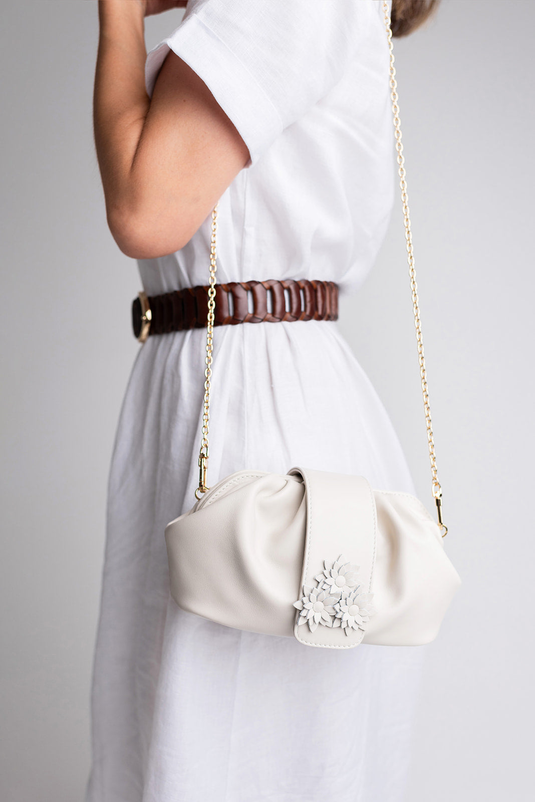 A cream-colored leather shoulder bag with a gold chain strap and a silver floral embellishment, worn crossbody by a model in a white dress with a brown belt.