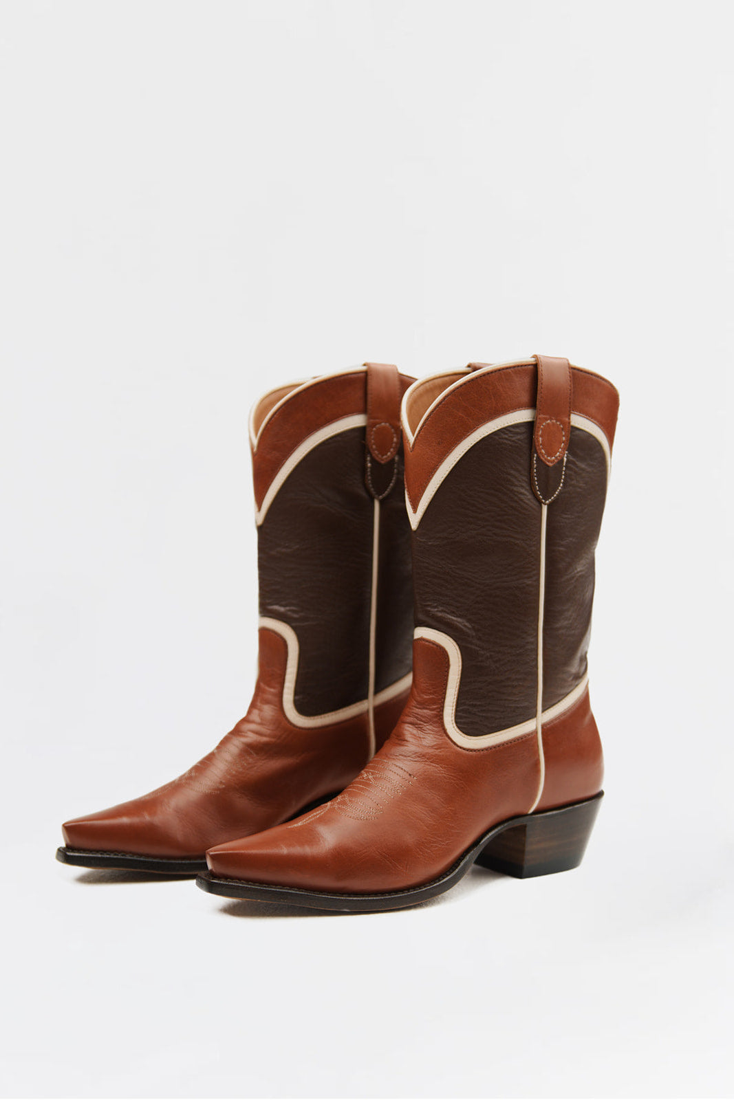 A pair of brown leather cowboy boots with dark brown shafts, cream-colored stitching, and low black heels, shown on a plain light gray background.