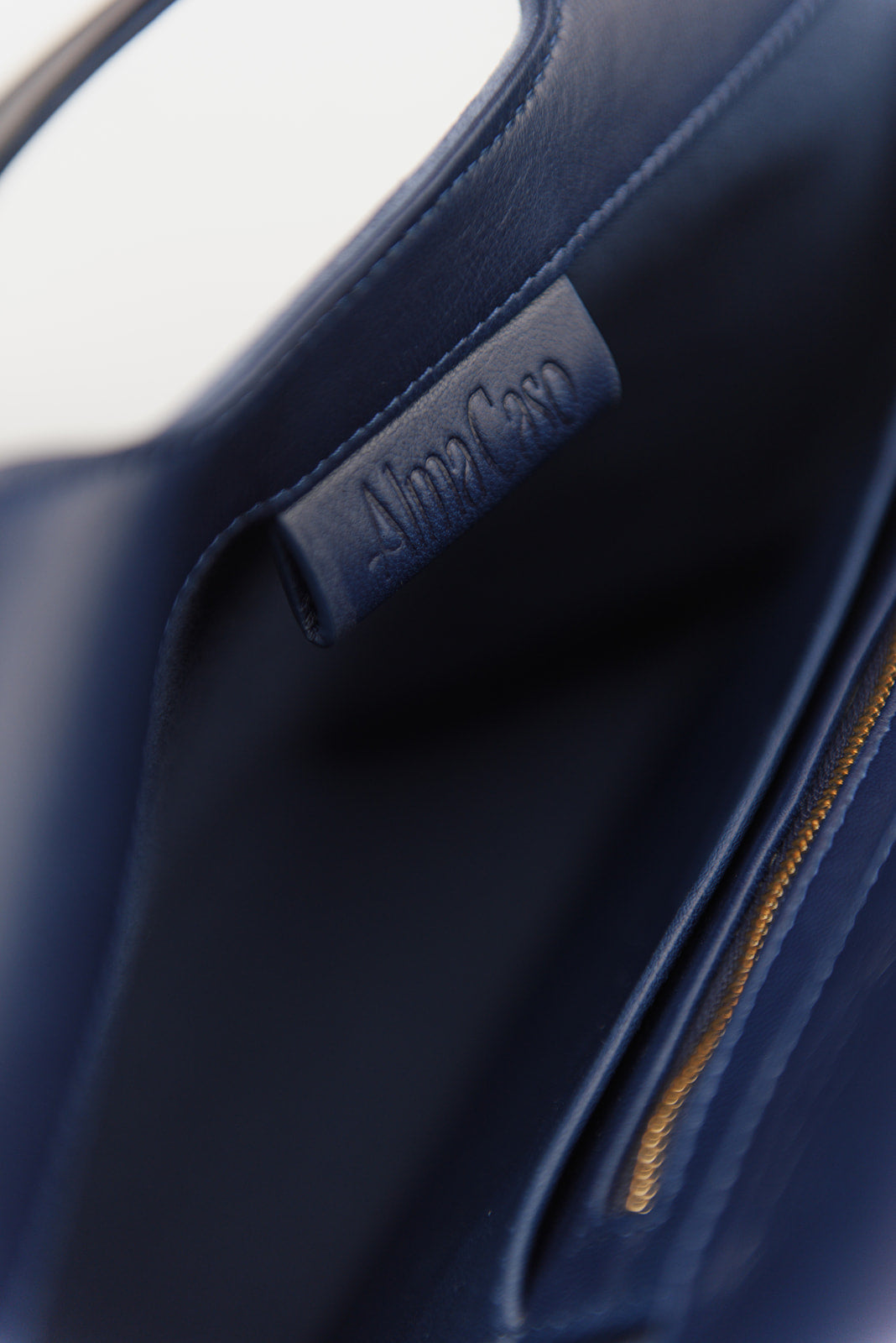 Close-up of a navy blue leather handbag interior showing a stitched brand label that reads Alma Cato and a gold zipper.