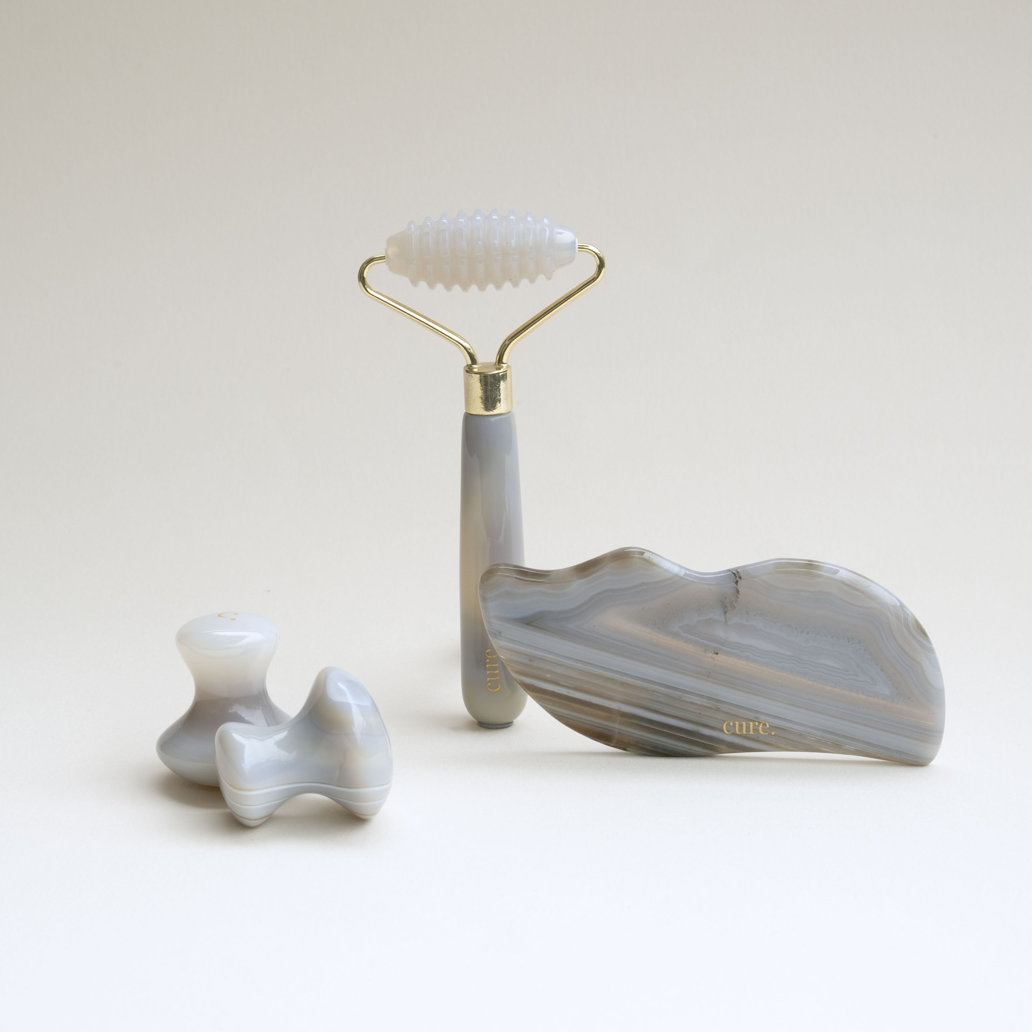 A facial massage set featuring a dual-ended roller with a white silicone head and a gray agate handle, a large leaf-shaped gua sha with gray and white agate bands, and two small wavy massage stones, all on a light beige background.
