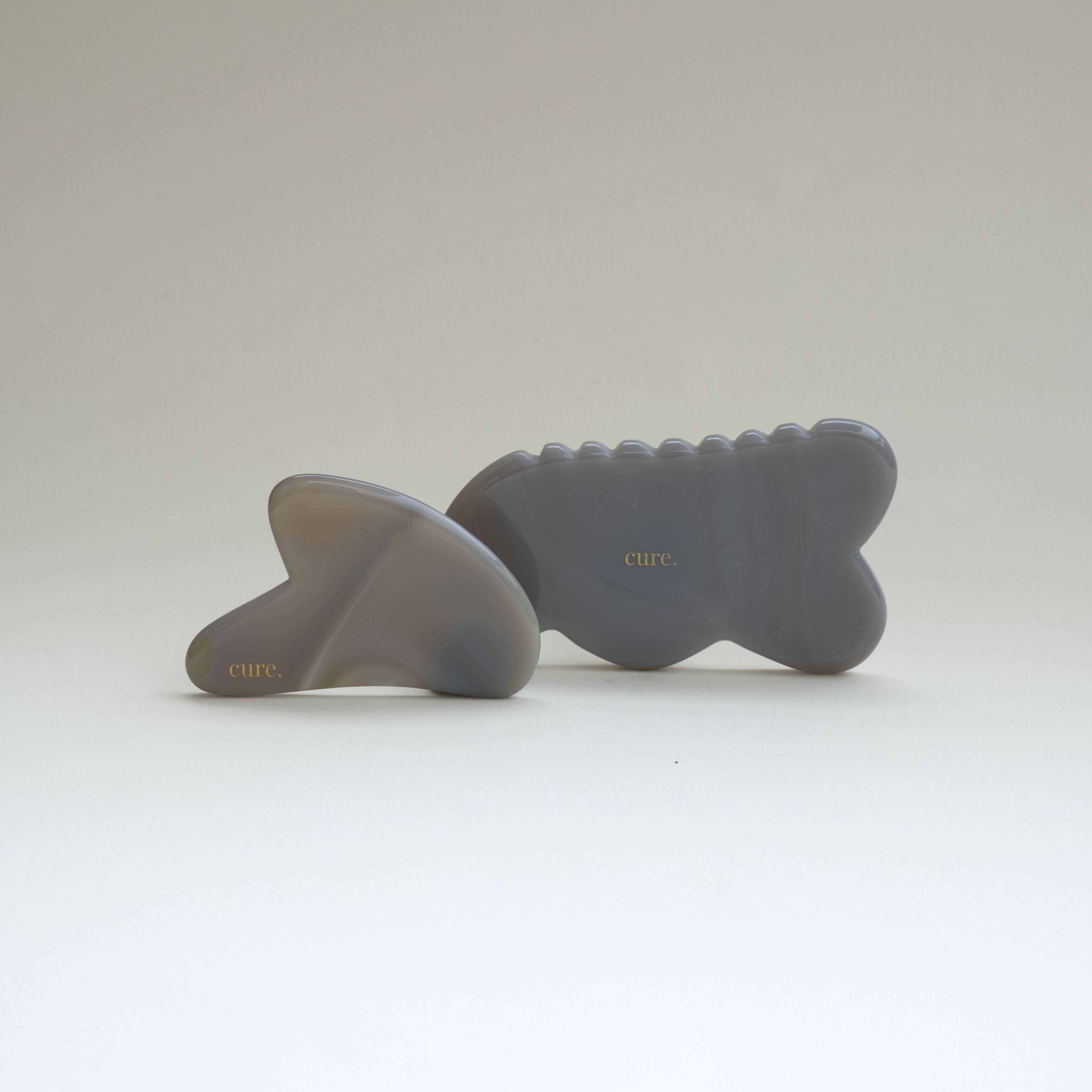 Two gray silicone massage tools with a wavy, fish-like silhouette and the brand name 'cure.' printed in gold on the front, displayed on a clean white studio background.