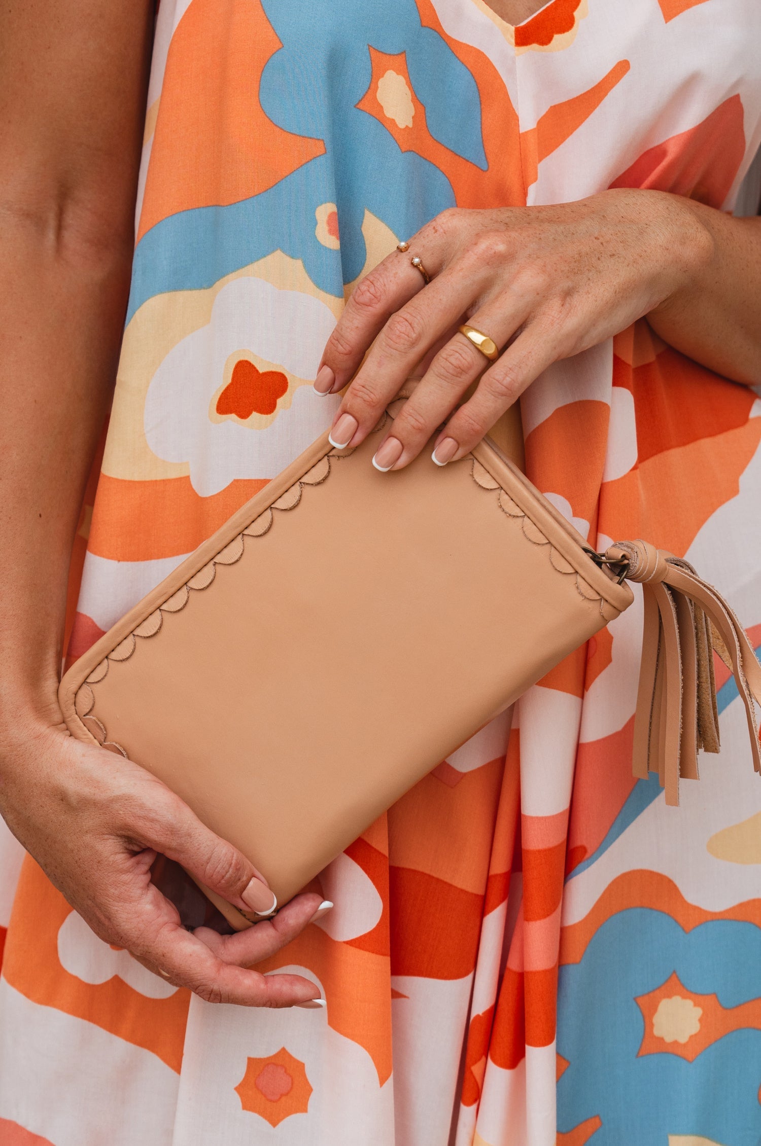 Person holding a tan clutch with a colorful floral dress in the background