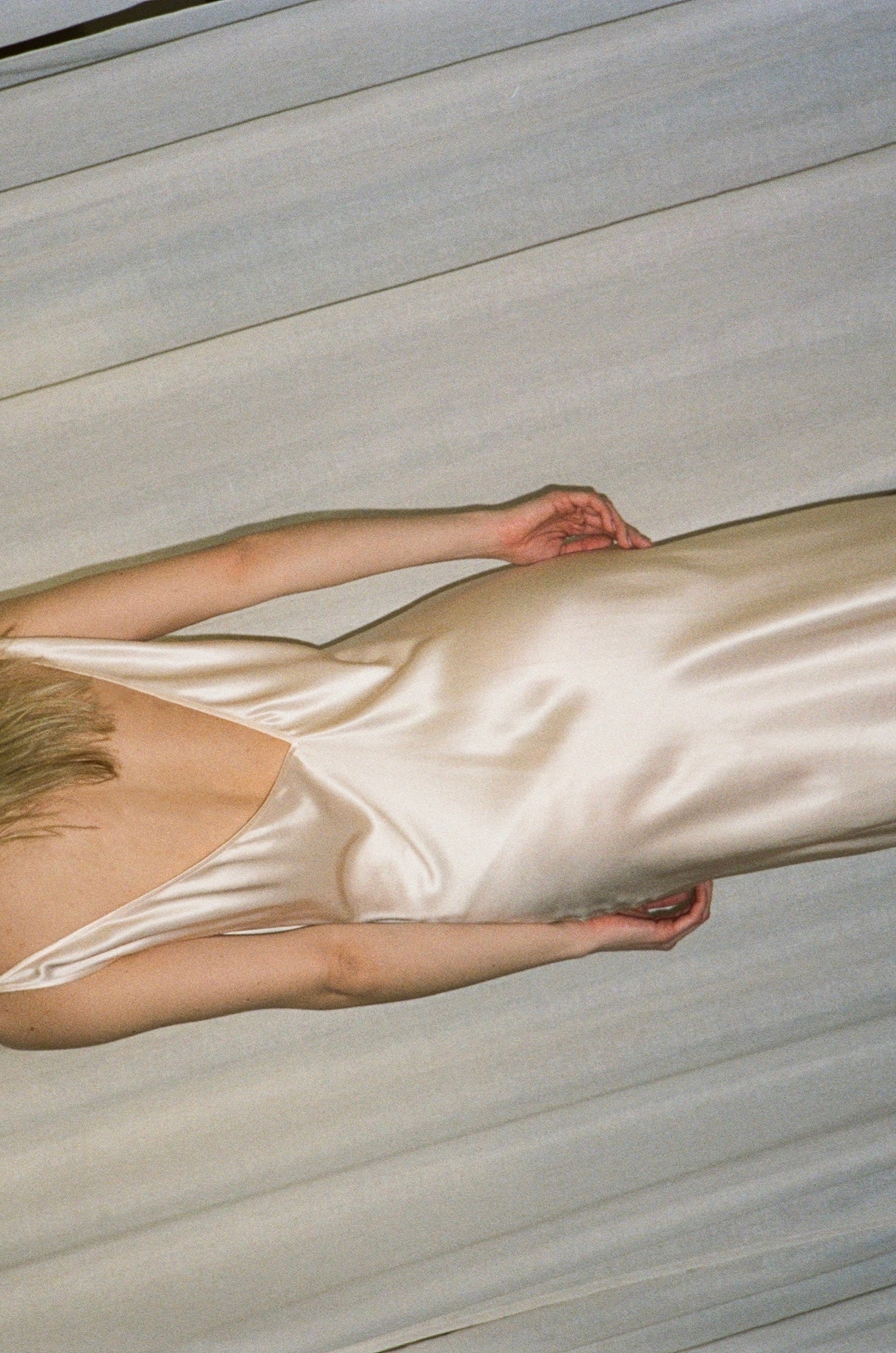 A model wearing a silk satin slip dress in a warm cream or champagne color, photographed from a high-angle perspective against a neutral, light gray textured background.