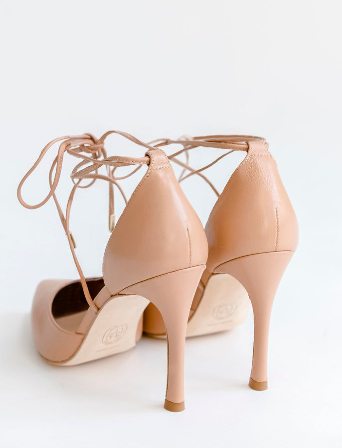 A pair of nude beige stiletto heels with lace-up ankle straps, shown from a rear three-quarter angle on a clean white background.