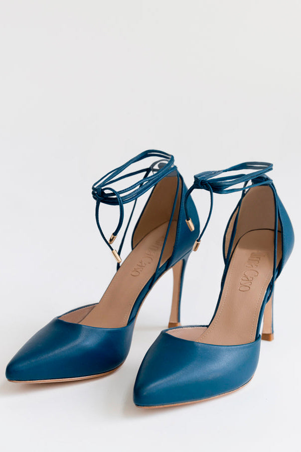 A pair of blue leather pointed-toe high heels with thin wraparound ankle straps and gold-tone hardware, photographed on a clean white studio background.