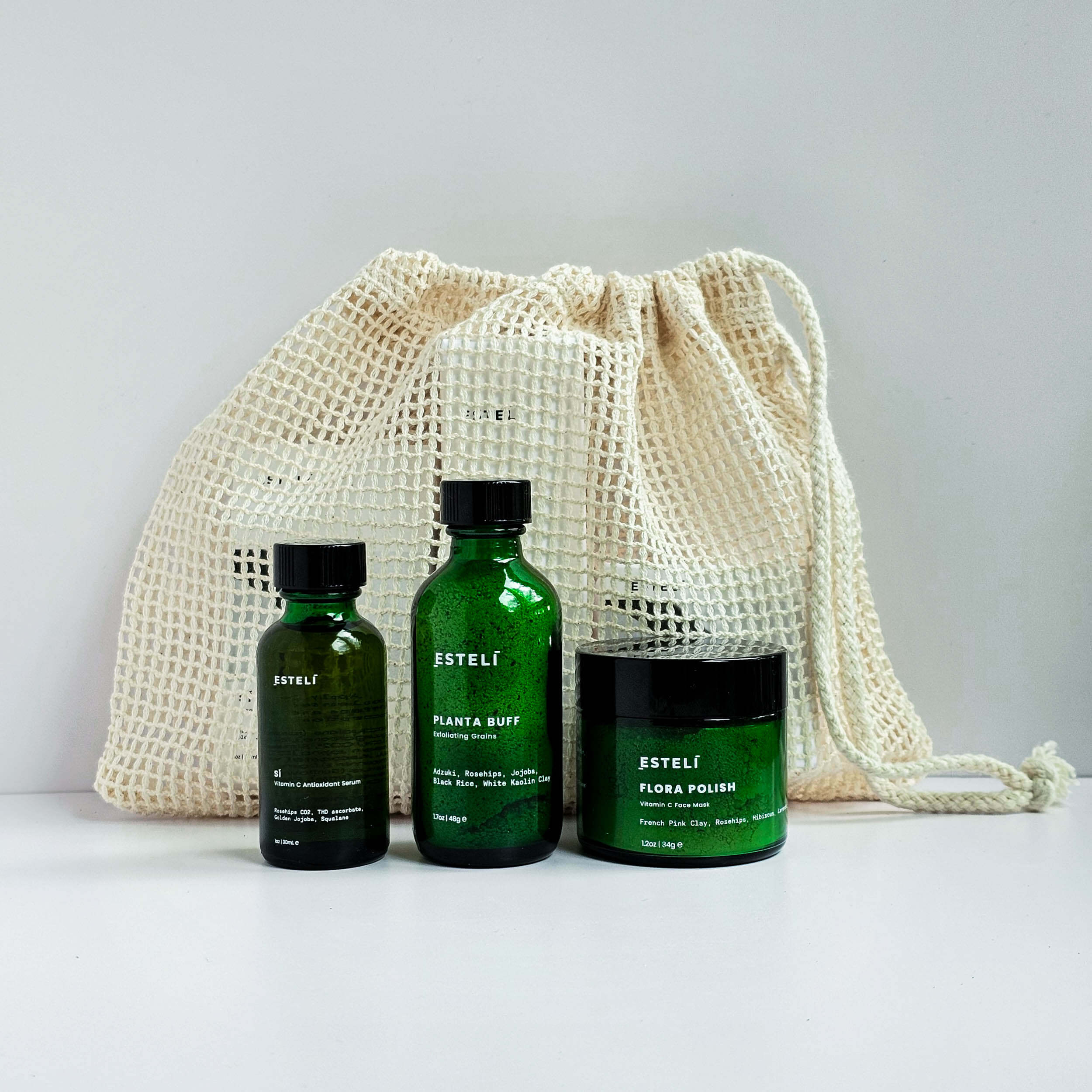 Three green bottles with black labels on a white surface next to a beige drawstring bag.