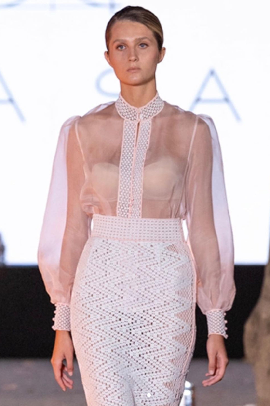 Model wearing a sheer pink top and white patterned skirt on a runway with a blurred background.