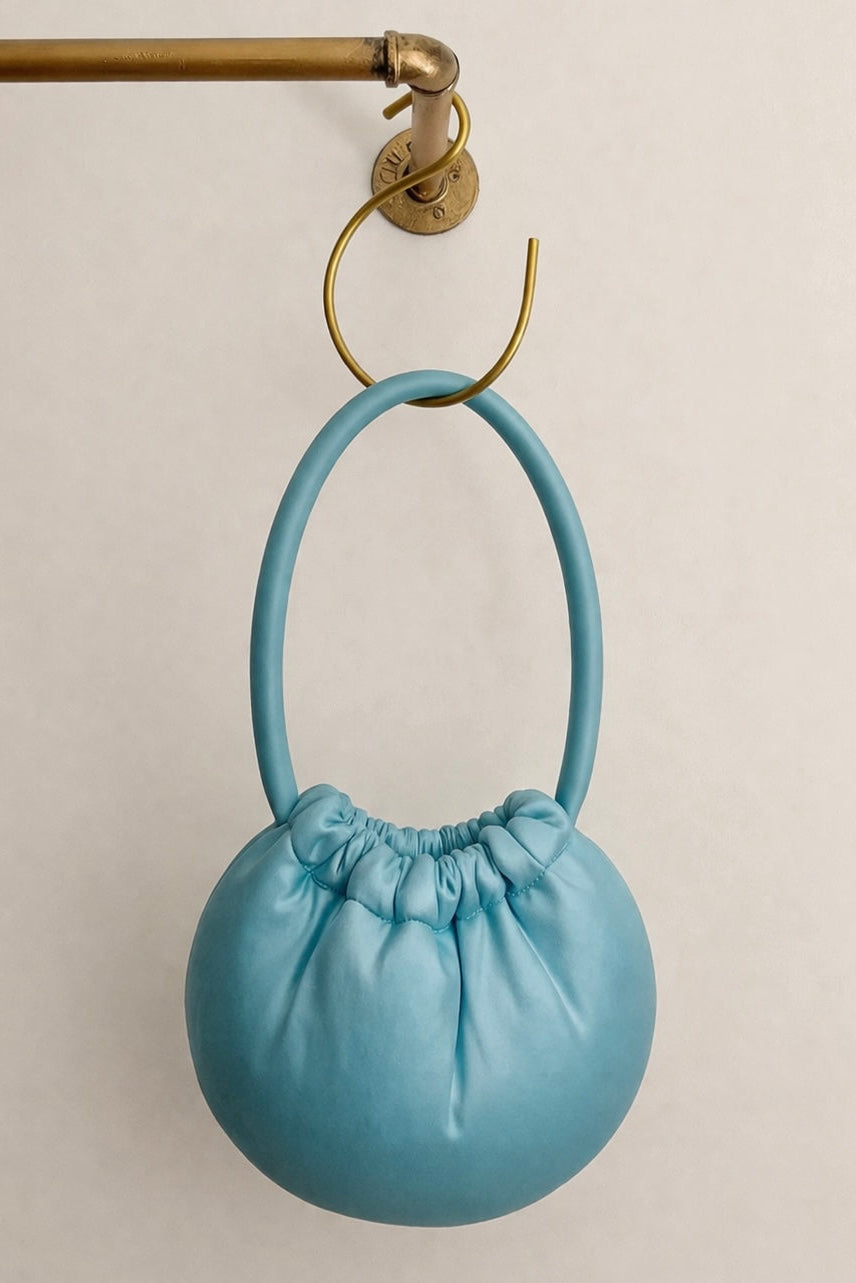 Light blue handbag with a round handle hanging on a gold hook against a beige background