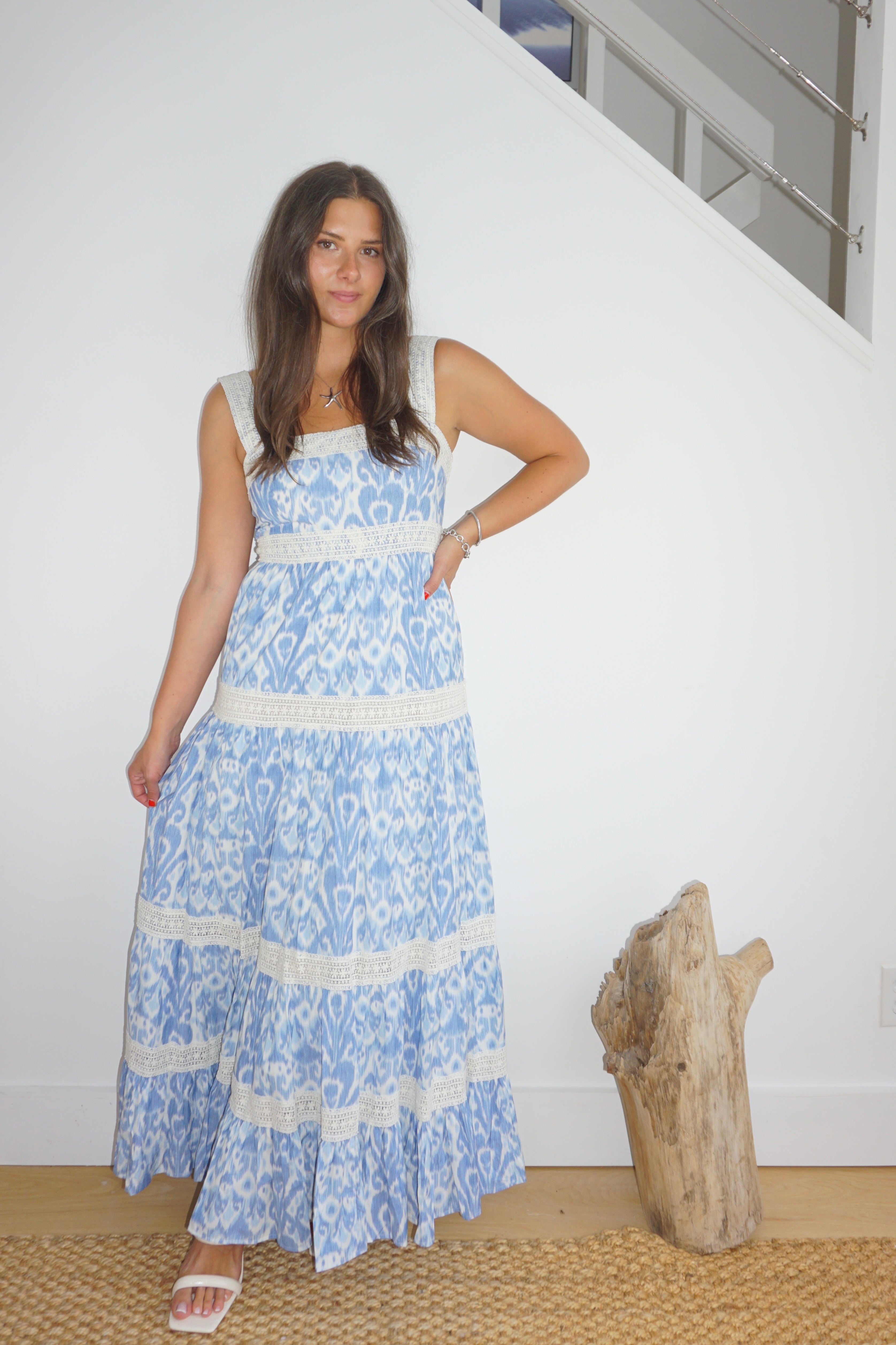 Woman wearing a blue floral dress standing indoors with a white wall and wooden object in the background.
