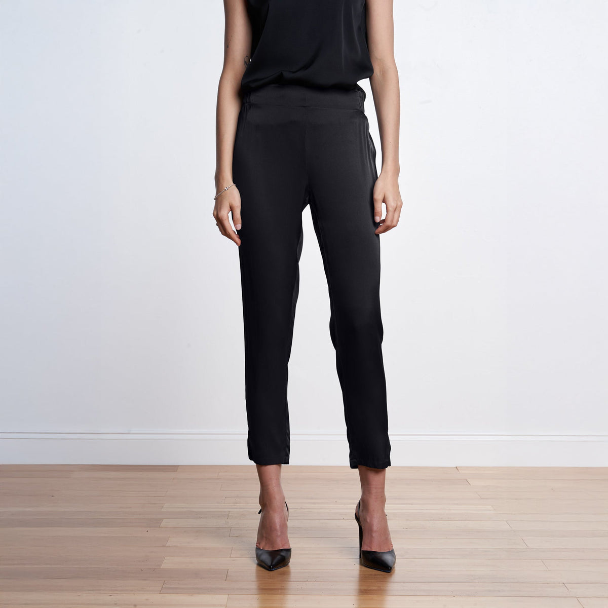 A model from the shoulders down wearing black cropped ankle-length silk trousers with side pockets, paired with a black sleeveless top and black pointed-toe heels, standing on a light wood floor against a white wall.
