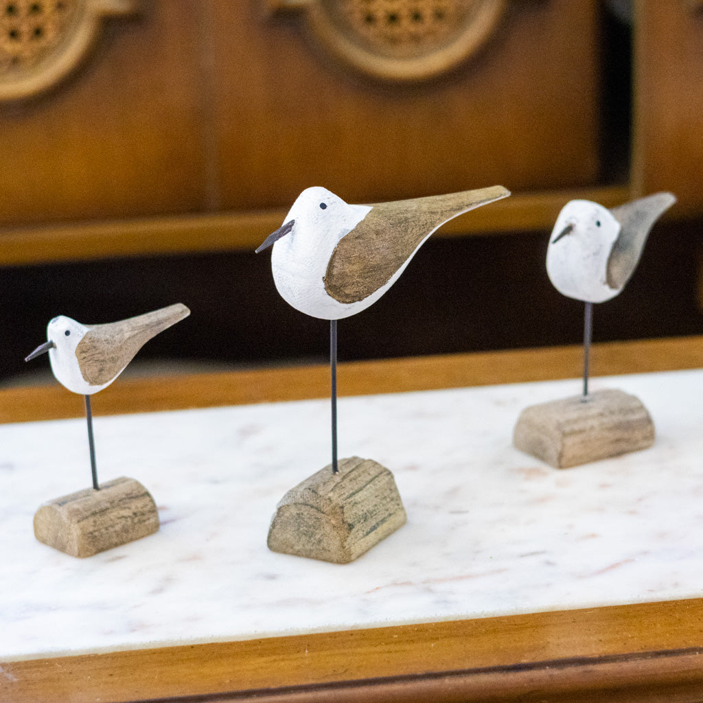 Three small bird figurines with white heads and brown wings, mounted on thin black wires and rustic wooden bases, arranged on a white marble surface with a warm wooden background.