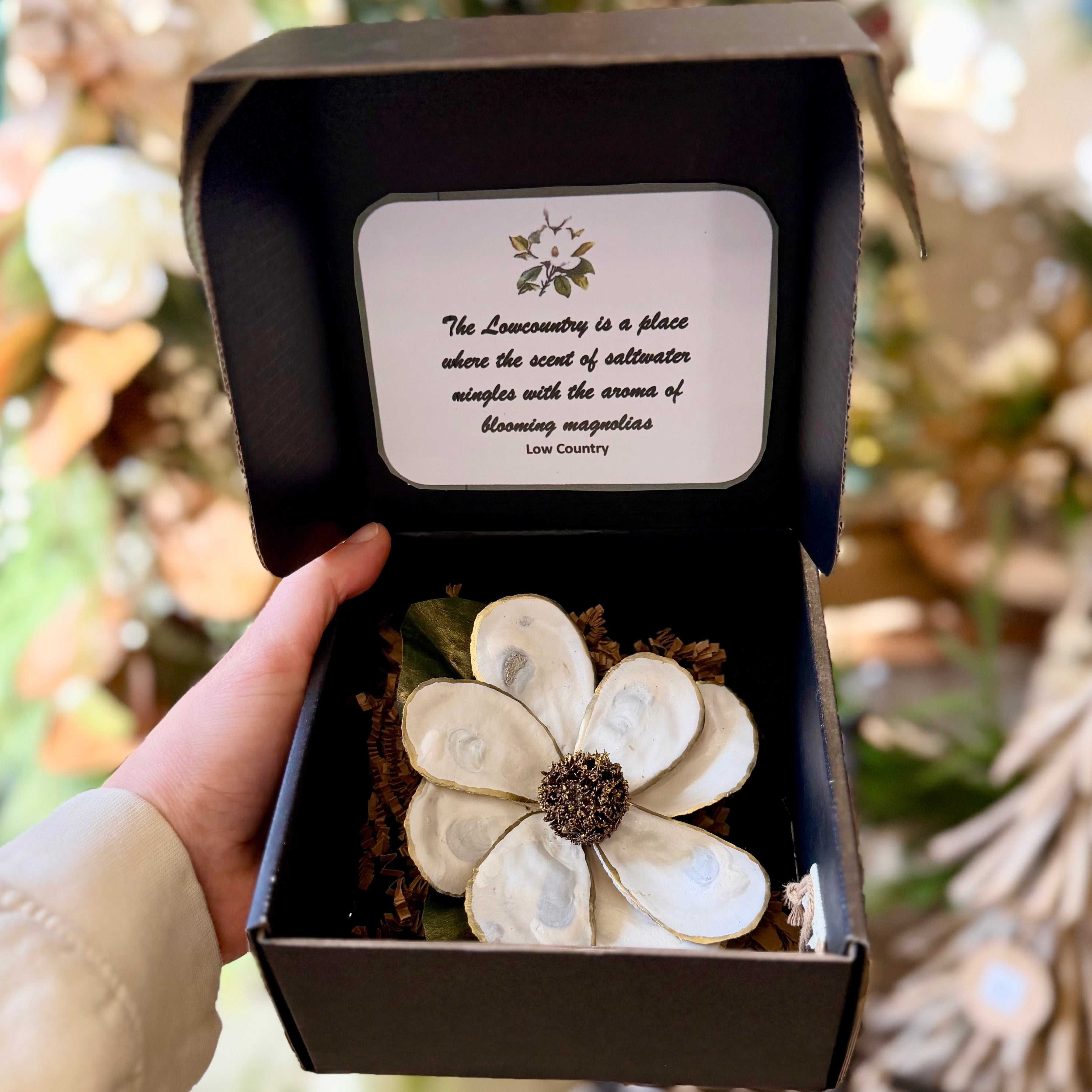 Hand holding a gift box with a white flower and text inside, against a blurred natural background.