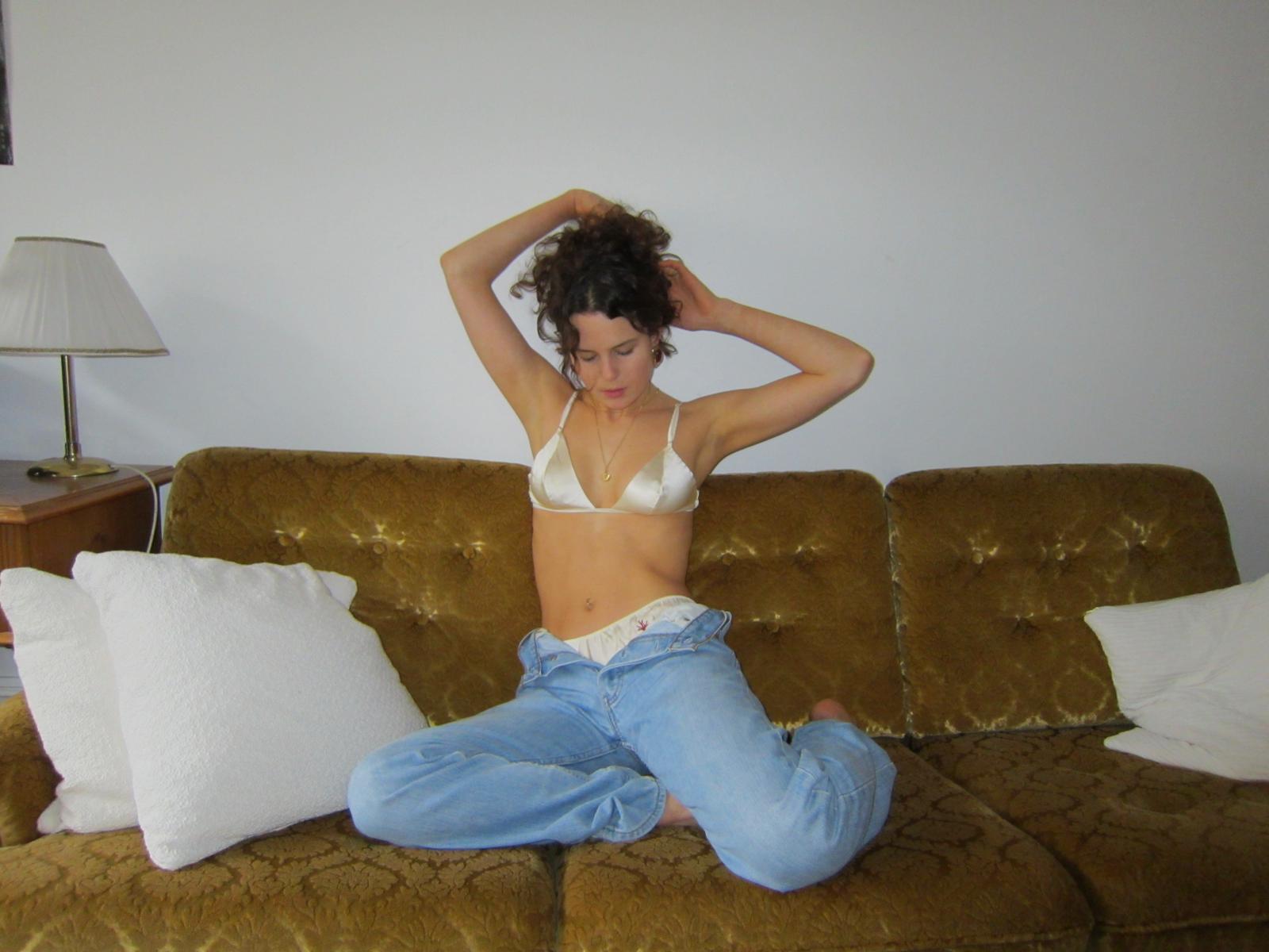 Woman sitting on a mustard colored retro couch wearing a silk bra, silk thong, and blue jeans.