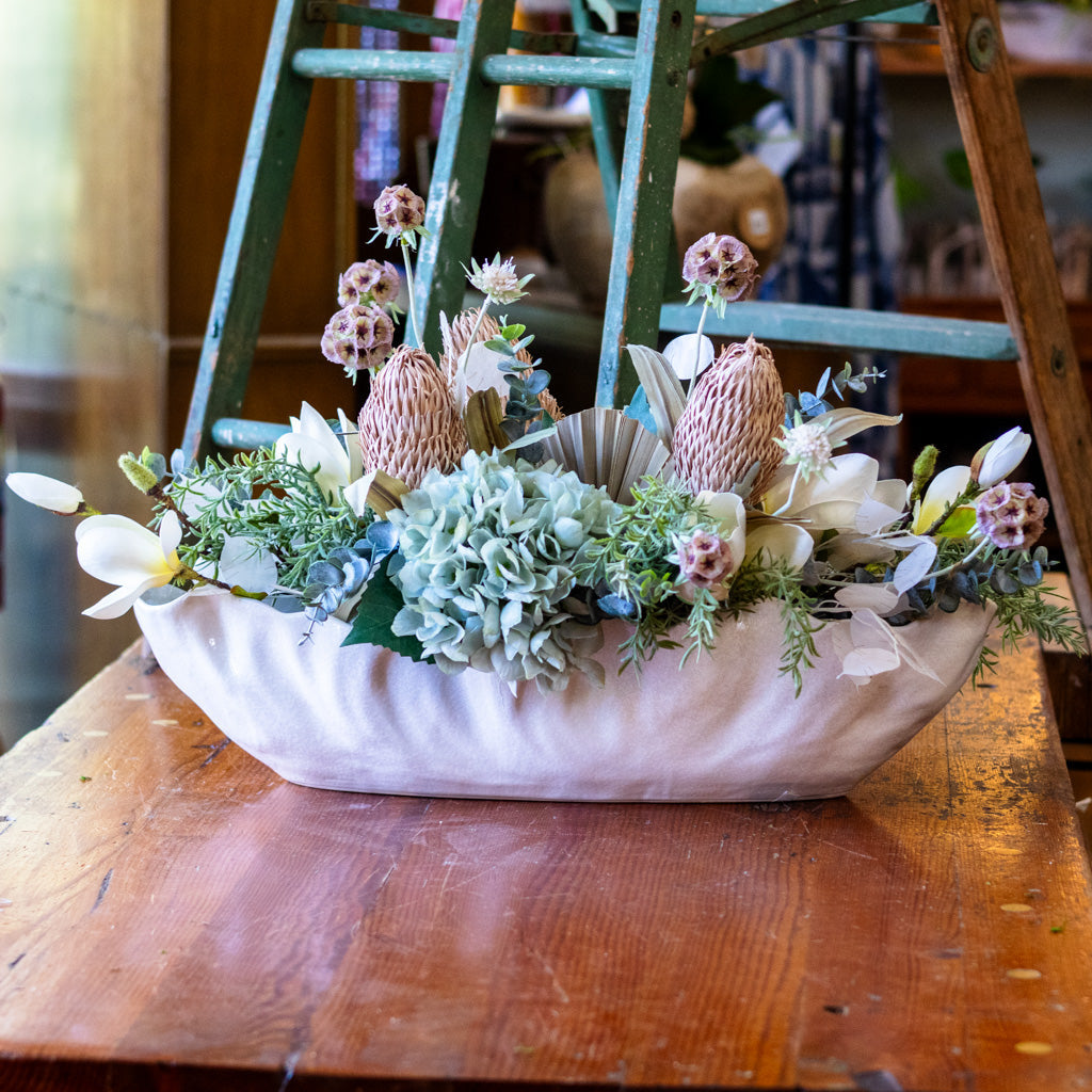 A decorative floral arrangement in a pale pink ceramic bowl, placed on a rustic wooden table with a green wooden ladder in the background.