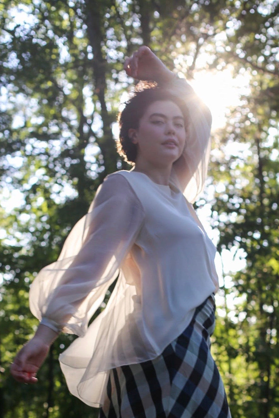 Woman in a white blouse and black and white striped skirt standing in a forest with sunlight filtering through the trees.