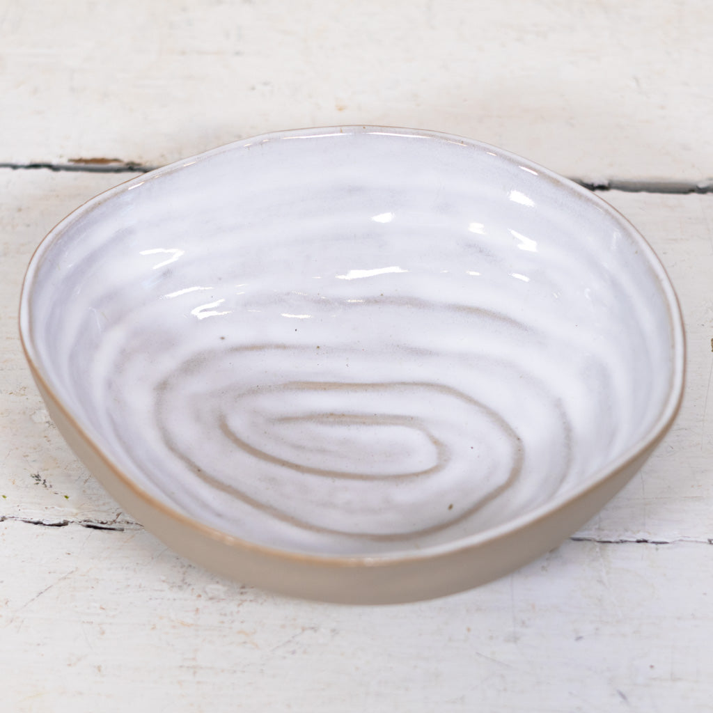 A shallow round shell-like ceramic bowl with a glossy white interior featuring subtle gray and beige concentric swirl patterns, and a matte beige exterior, set on a distressed white wooden surface.