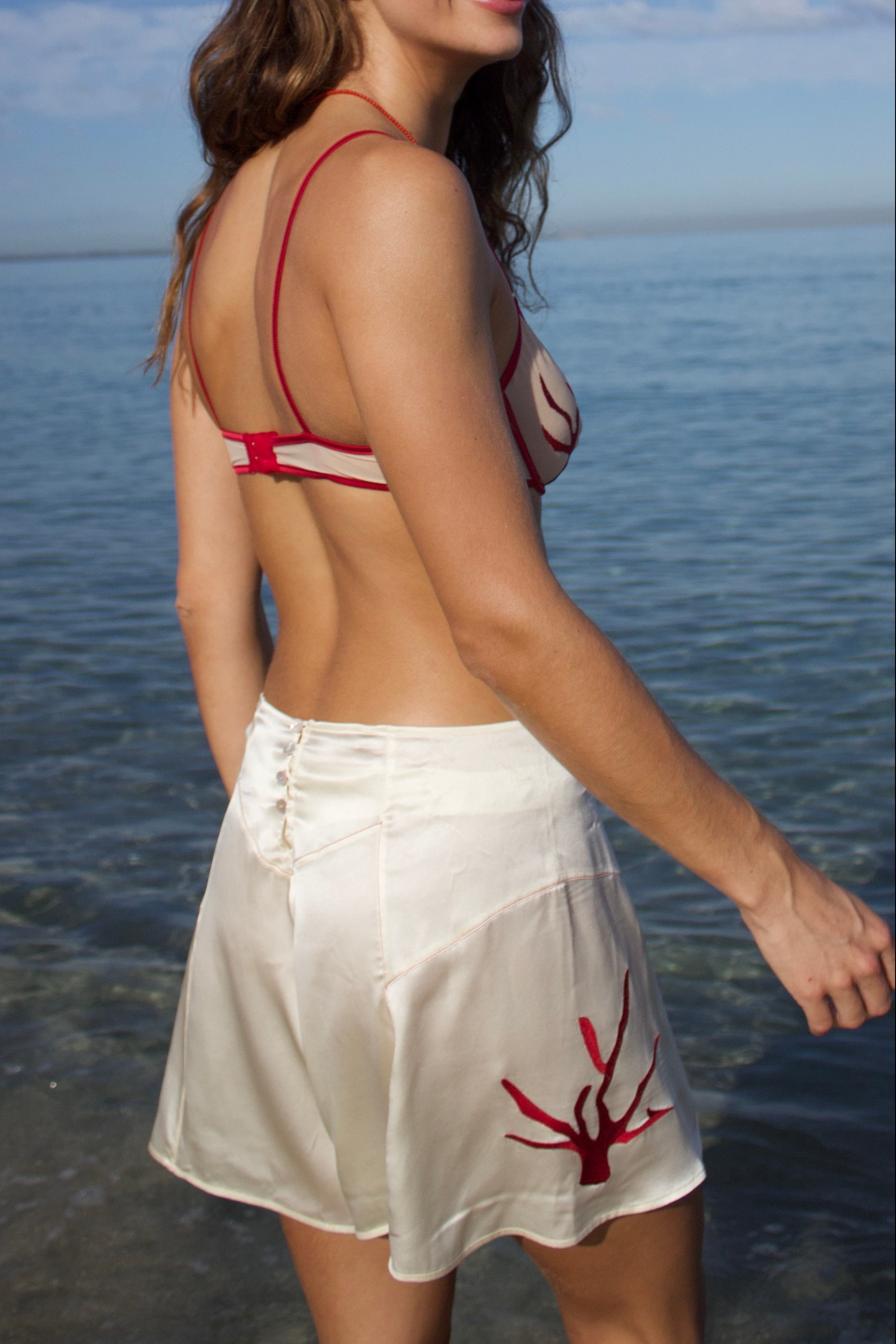 Woman wearing a beige and red coral bra with white silk shorts featuring a red coral design, standing by the water.