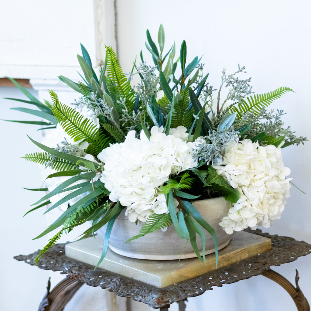 Floral arrangement with white flowers and green leaves in a white vase on a decorative stand.