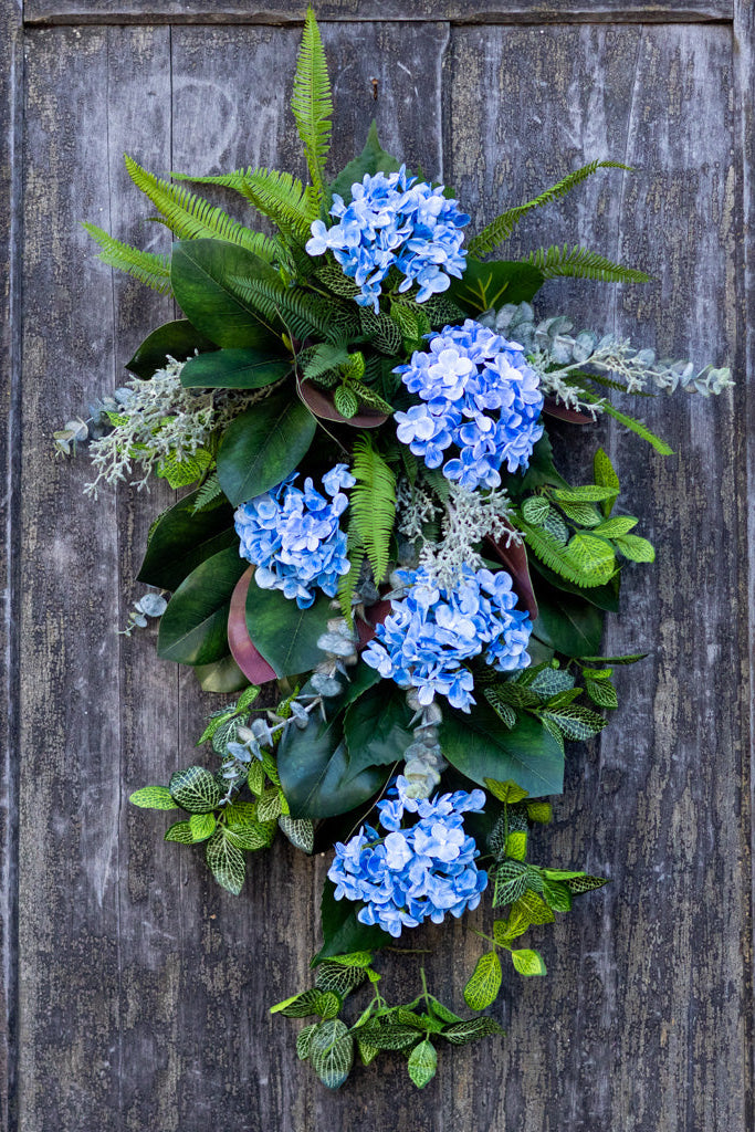 A decorative floral wreath made of blue hydrangea blooms, green ferns, and leafy greenery, hung on a weathered dark wood door.