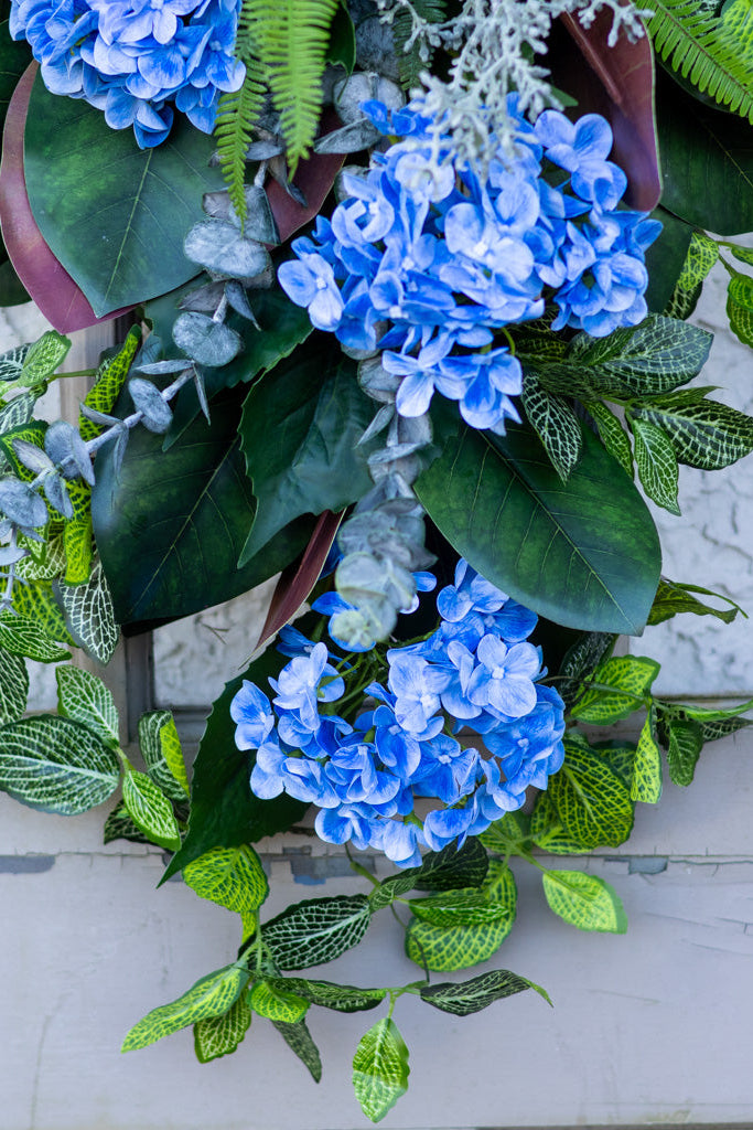 A hanging floral wreath featuring clusters of blue hydrangeas, large green leaves, and green-and-white patterned foliage, displayed against a light textured wall.